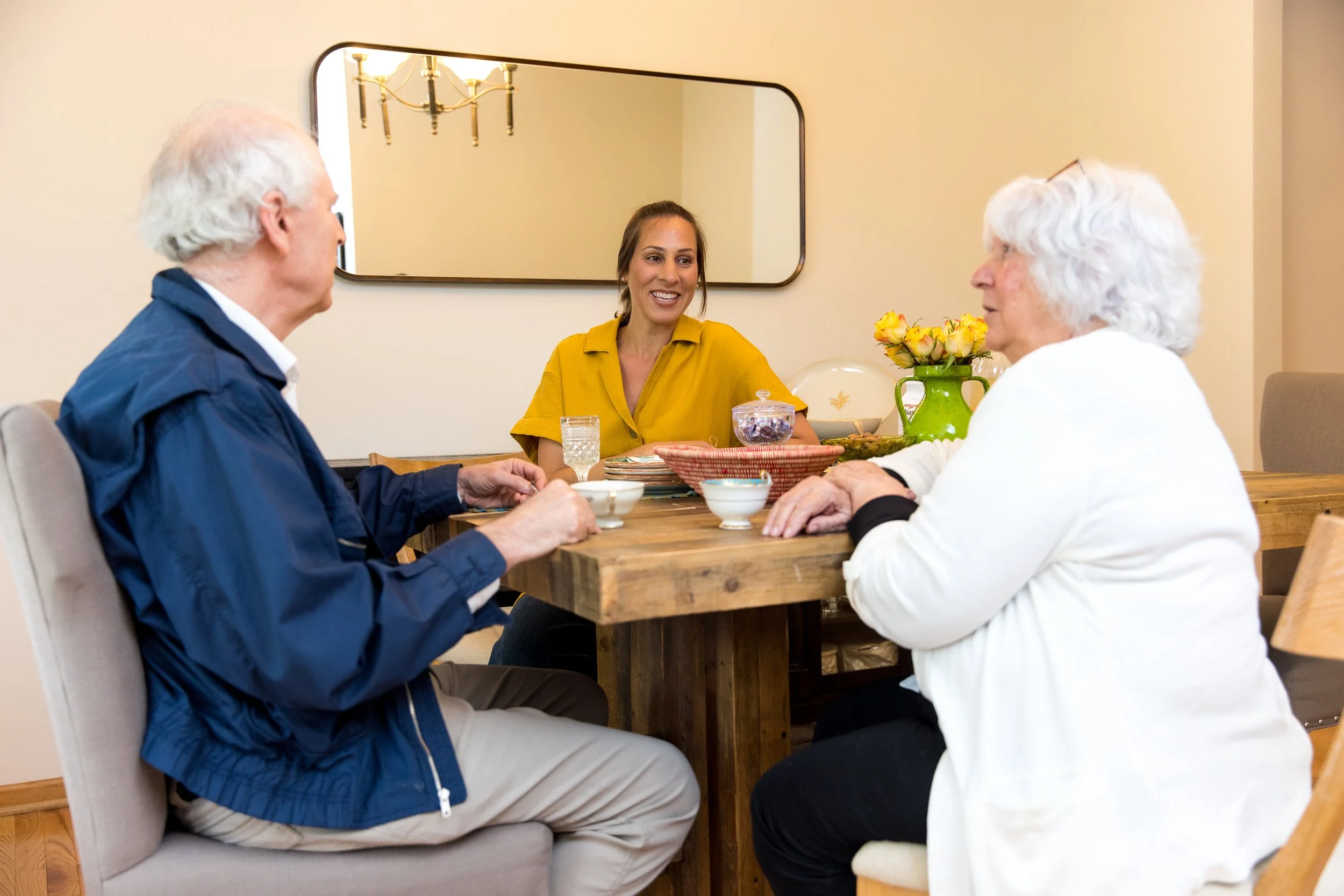 Three people sit around a wooden dining table having a conversation. The scene includes a man with white hair in a blue jacket, a woman with white hair in a white sweater, and a smiling woman in a yellow shirt. The table has cups, bowls, a green vase with yellow flowers, and a glass of water.