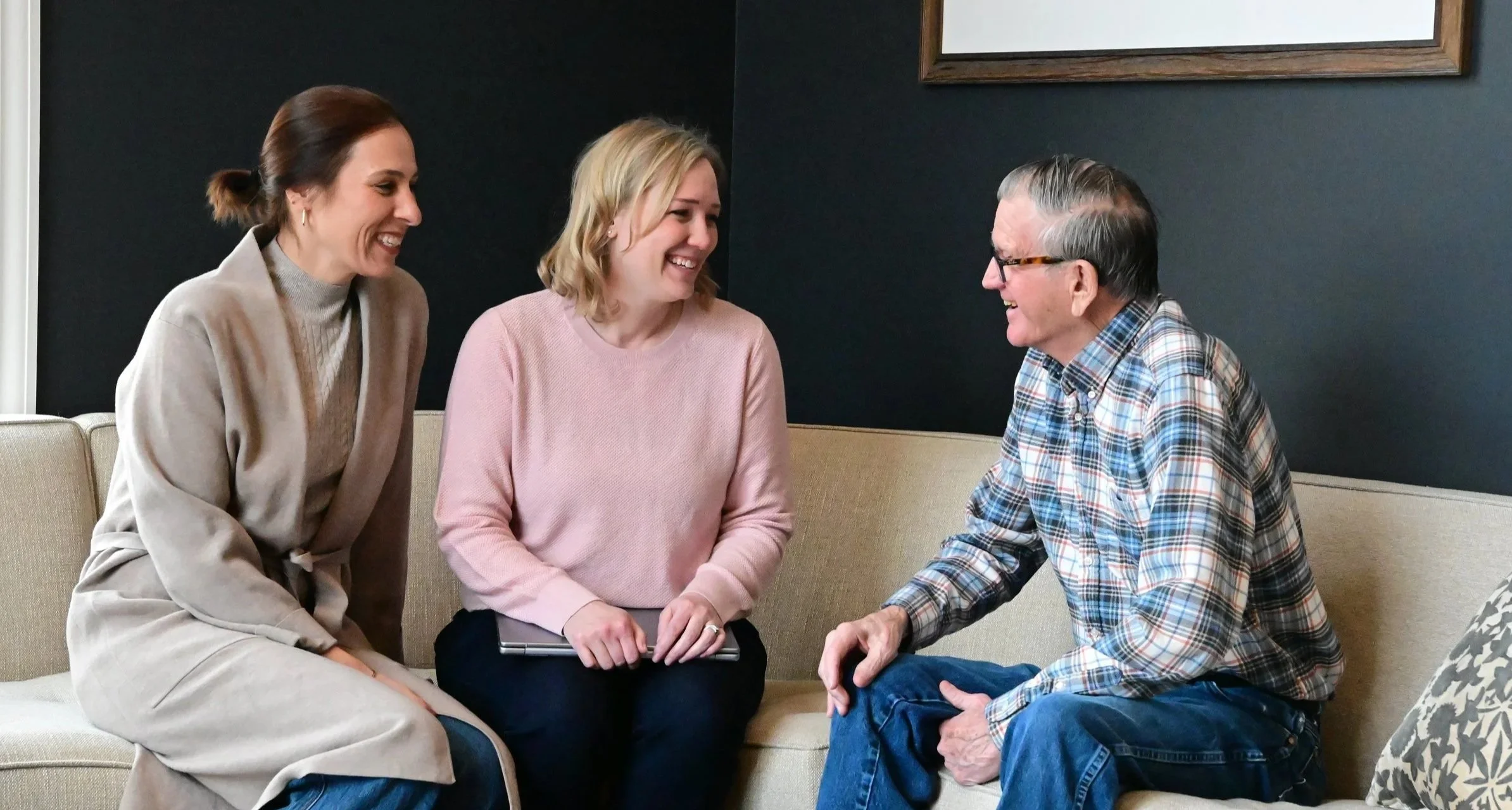 Three women and one older man sitting on a beige couch, smiling and engaging in conversation, in a room with dark walls.