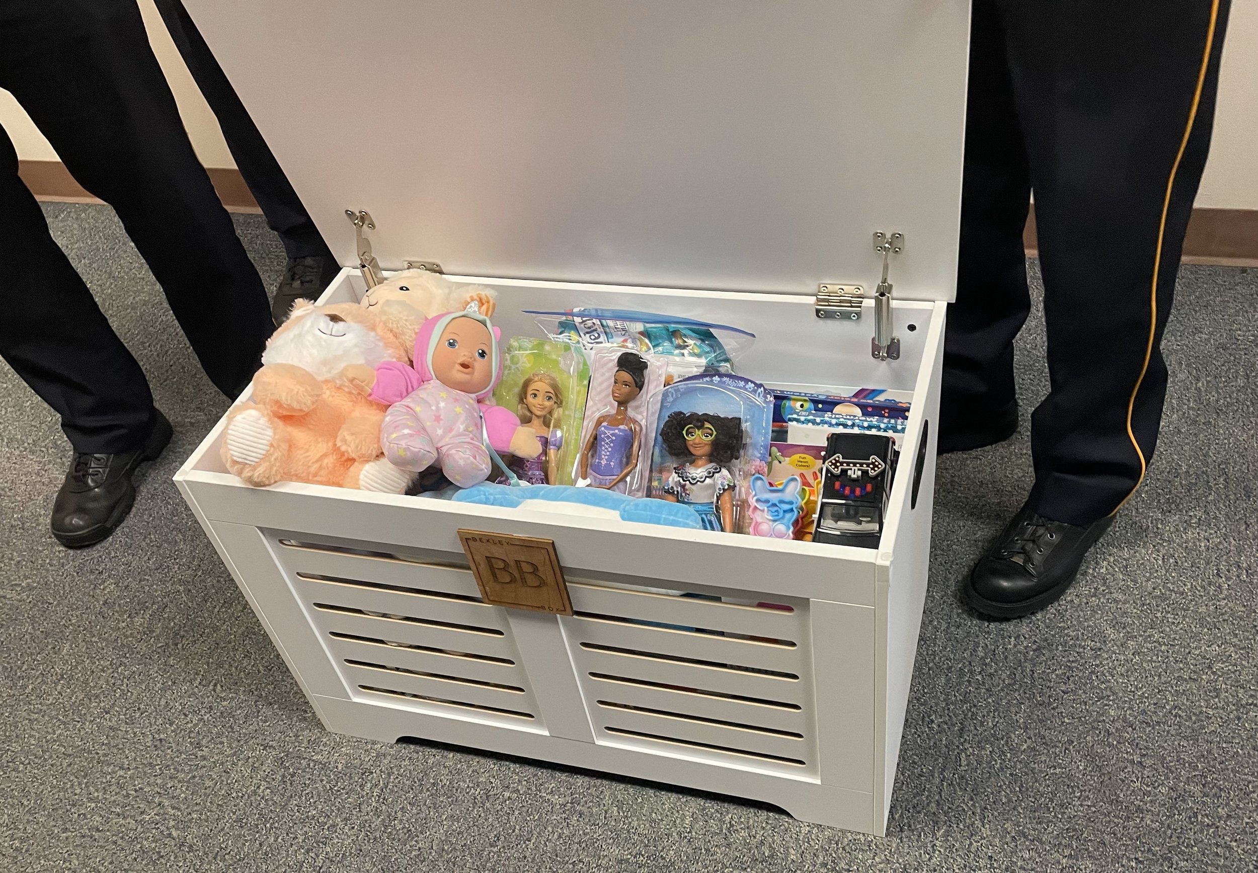 Open white toy chest filled with various toys including plush animals, dolls, and small games, with two people in black pants and shoes standing on either side.