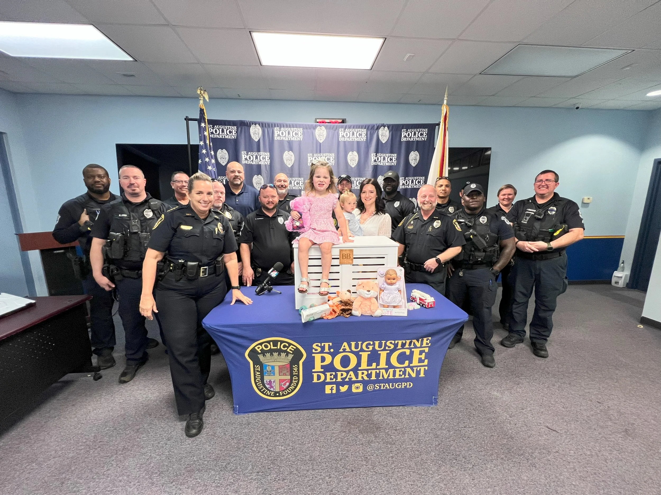 Group of police officers and children gathered around a table with toys, at the St. Augustine Police Department, celebrating a special event.