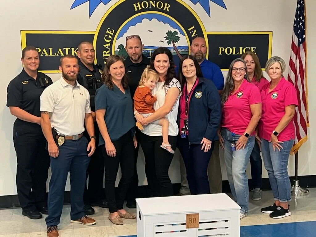 Group photo of police officers, city officials, and a woman holding a young girl, in front of a police department emblem. The group is smiling, with some in uniform and others in casual or pink shirts, standing indoors next to an American flag.