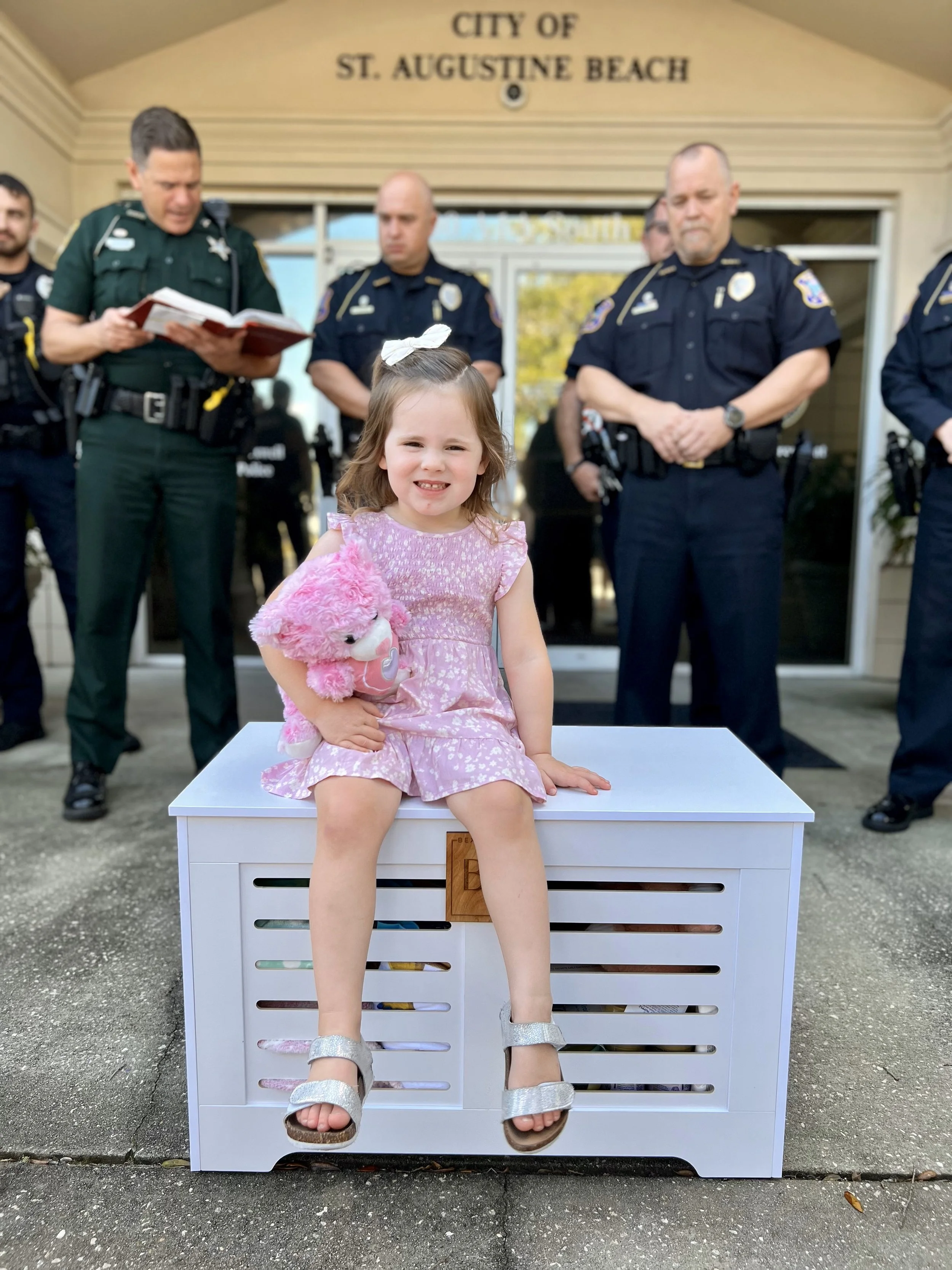 Young girl sitting on a white crate, holding a pink stuffed animal, smiling, with police officers and a sheriff standing behind her in front of the City of St. Augustine Beach building.