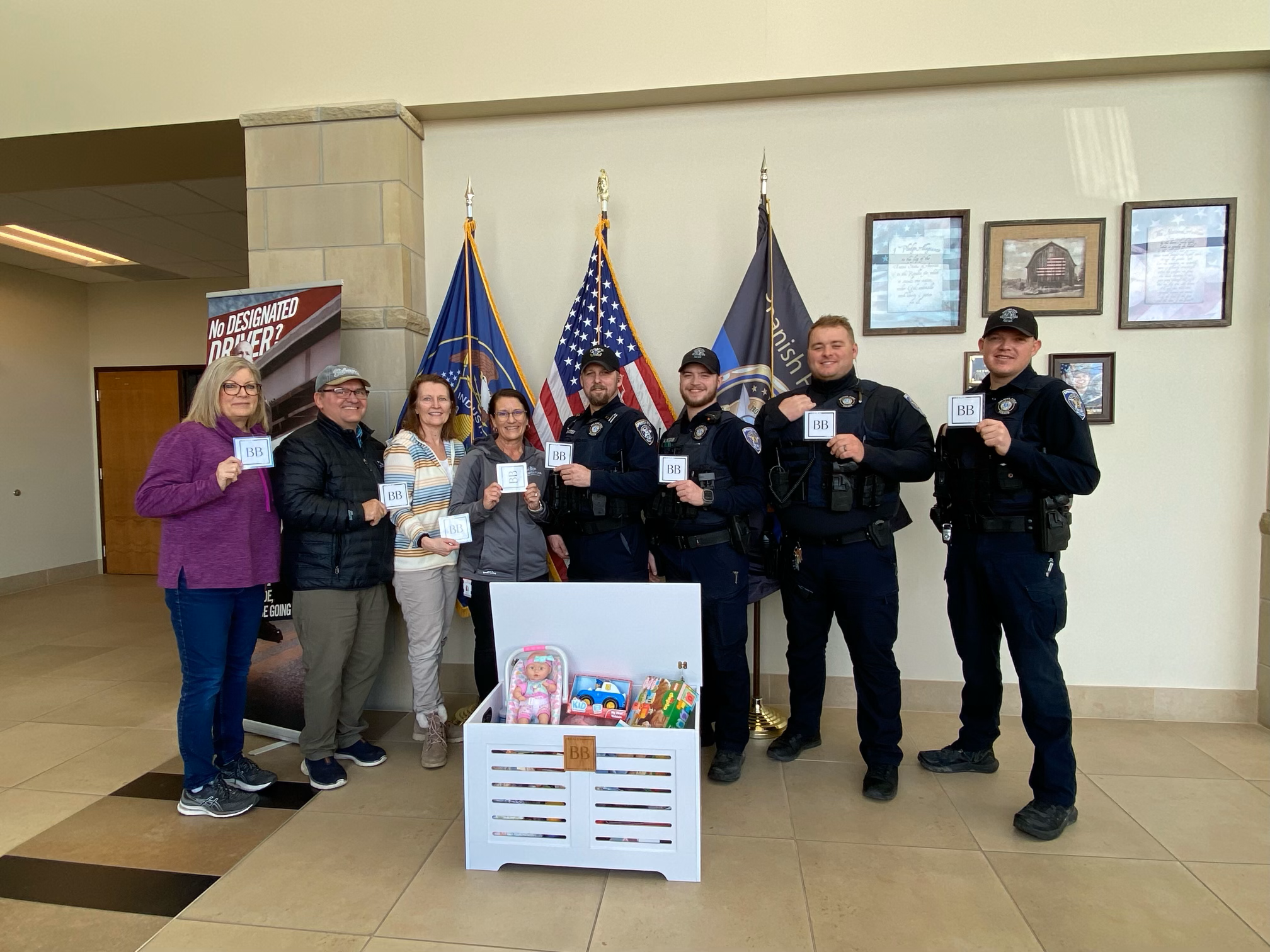 Group of women and police officers standing indoors, holding stickers, in front of flags and framed pictures, with a donation box containing toys at their feet.