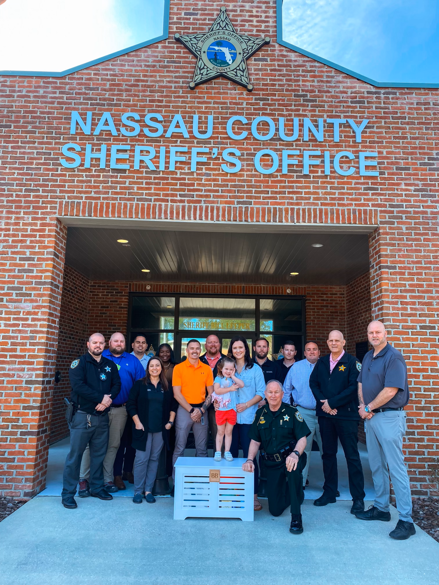 Group photo of people, including sheriff and officers, standing outside the Nassau County Sheriff's Office building with a young girl in front, near a white crate.