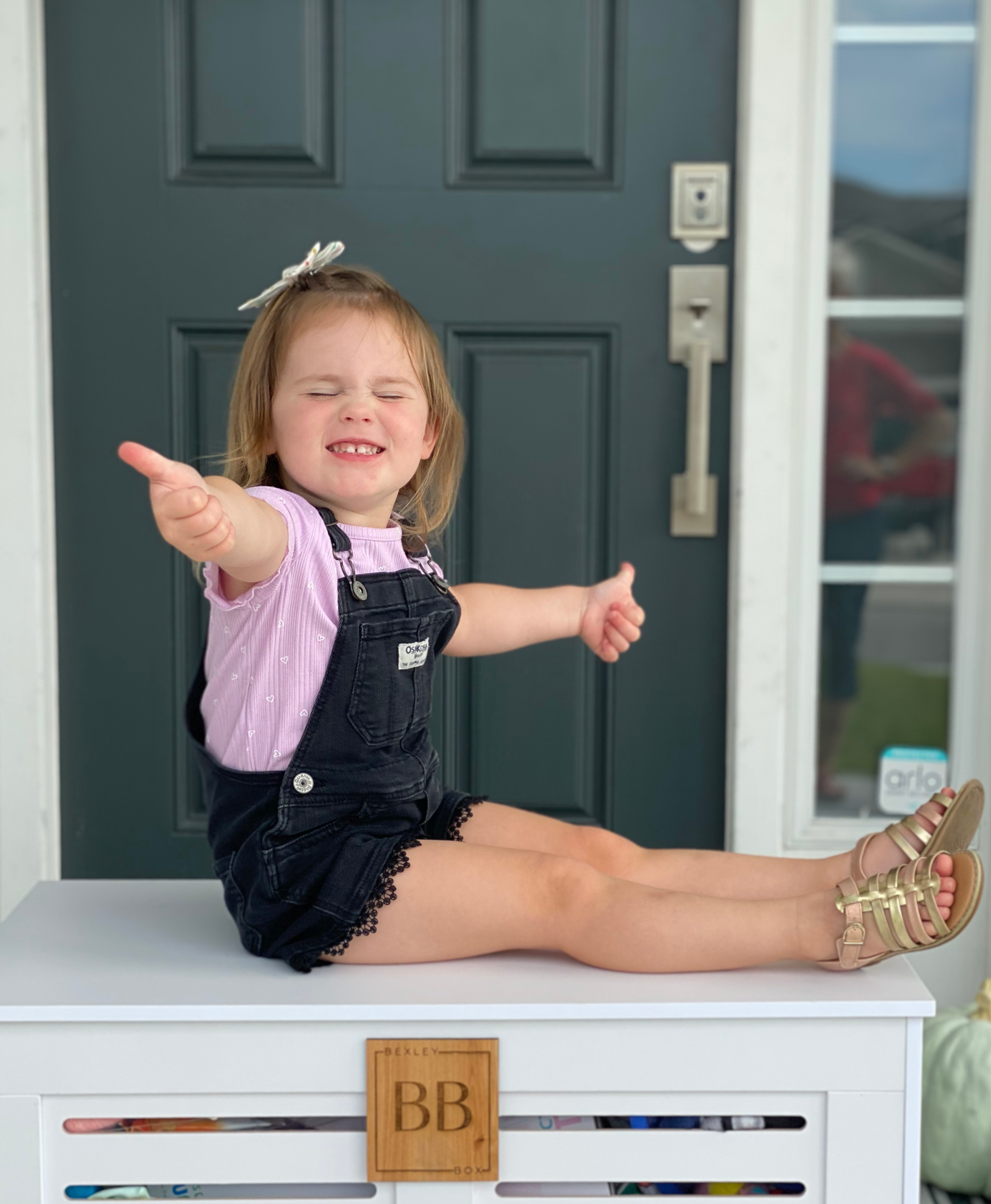 A young girl with a bow in her hair, wearing a pink shirt and black overalls, sitting on a white storage box on a porch, smiling and pointing outward with her eyes closed.