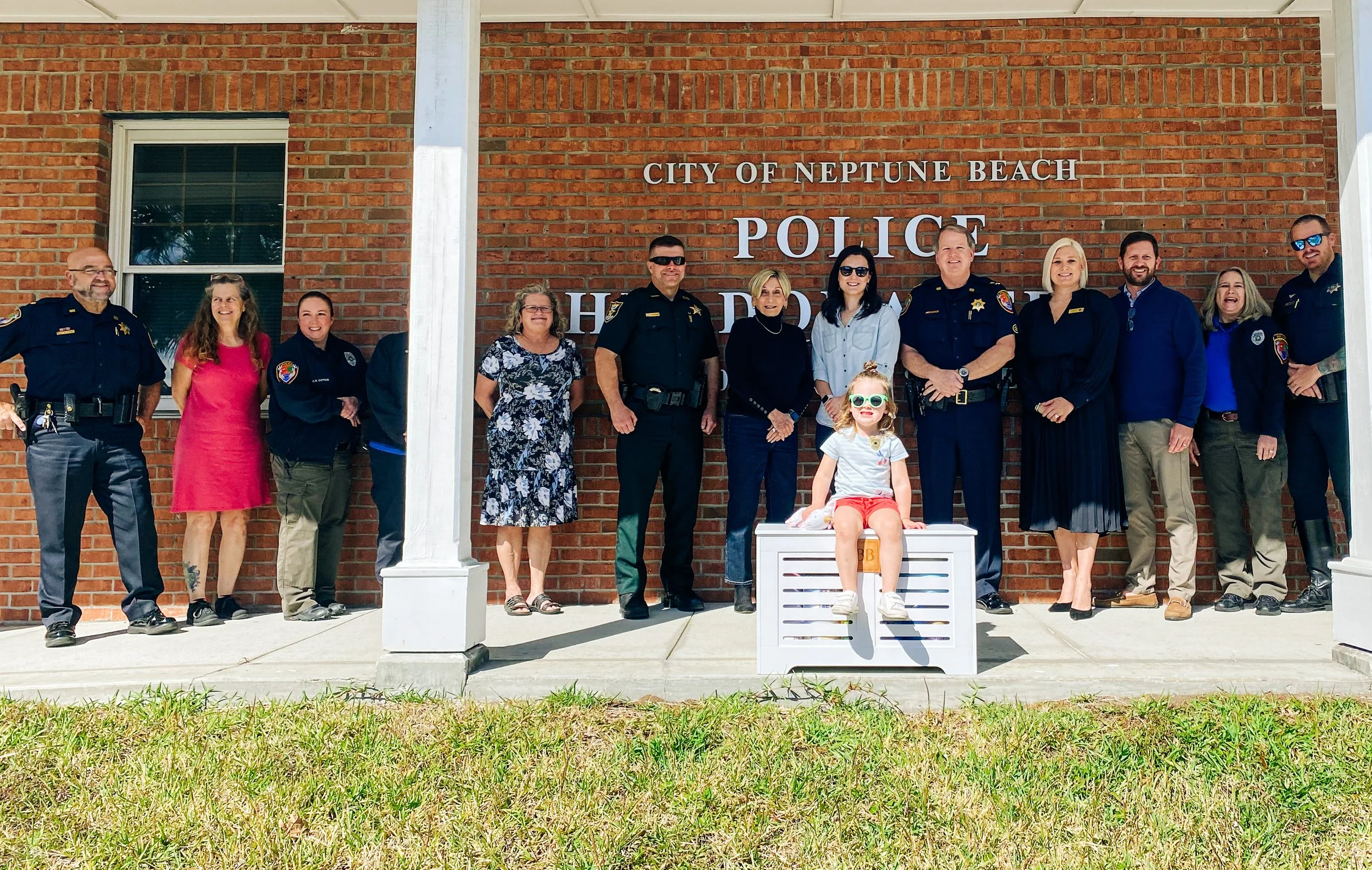 Group of police officers and civilians standing in front of the City of Neptune Beach Police Department building, with a young girl sitting on a white bench in the foreground.