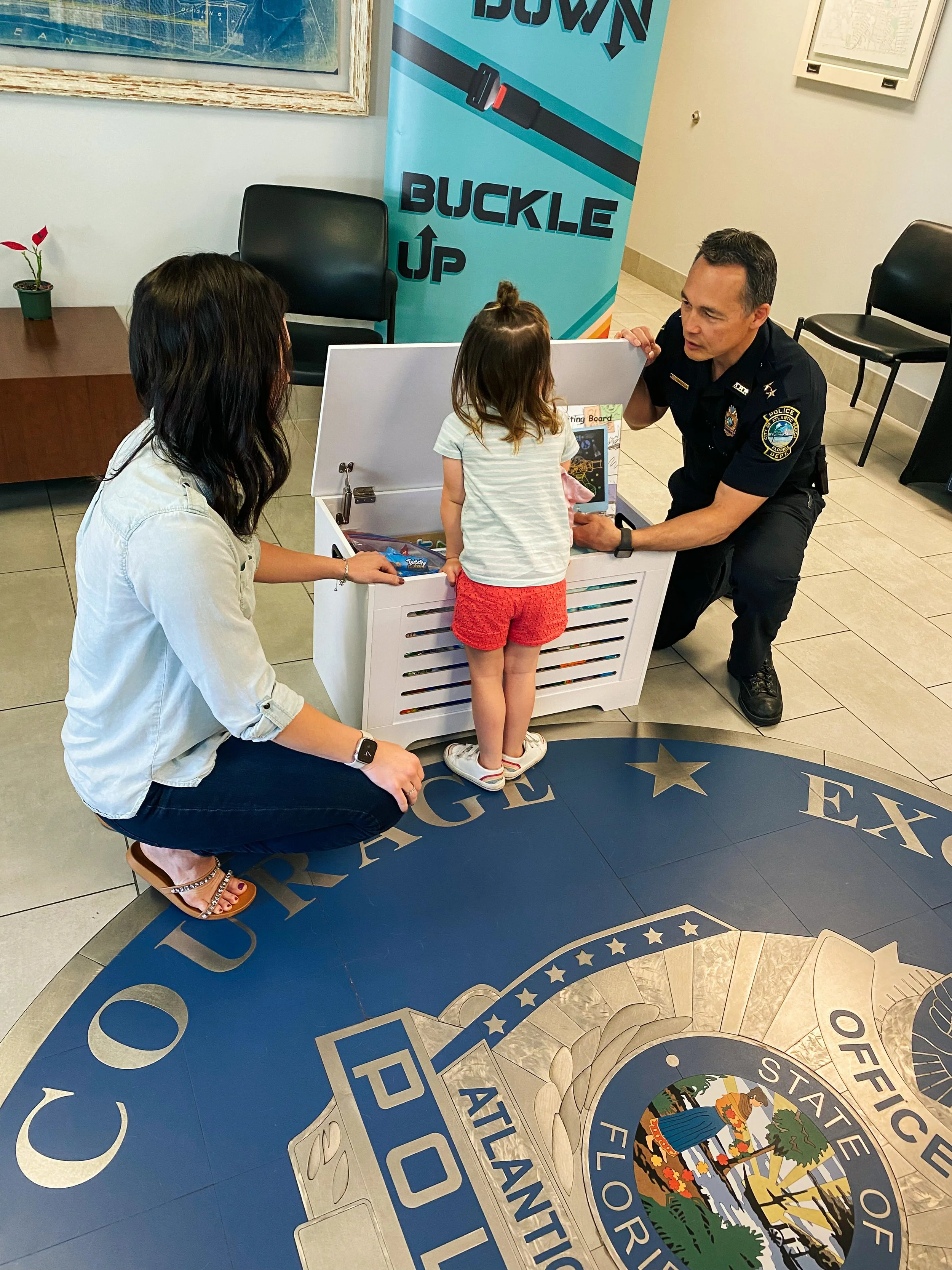 A young girl is talking to a police officer while a woman kneels nearby. They are in a room with the Florida state police seal on the floor. The girl is standing next to a white toy chest. A police badge and a police patch are visible. A large poster
