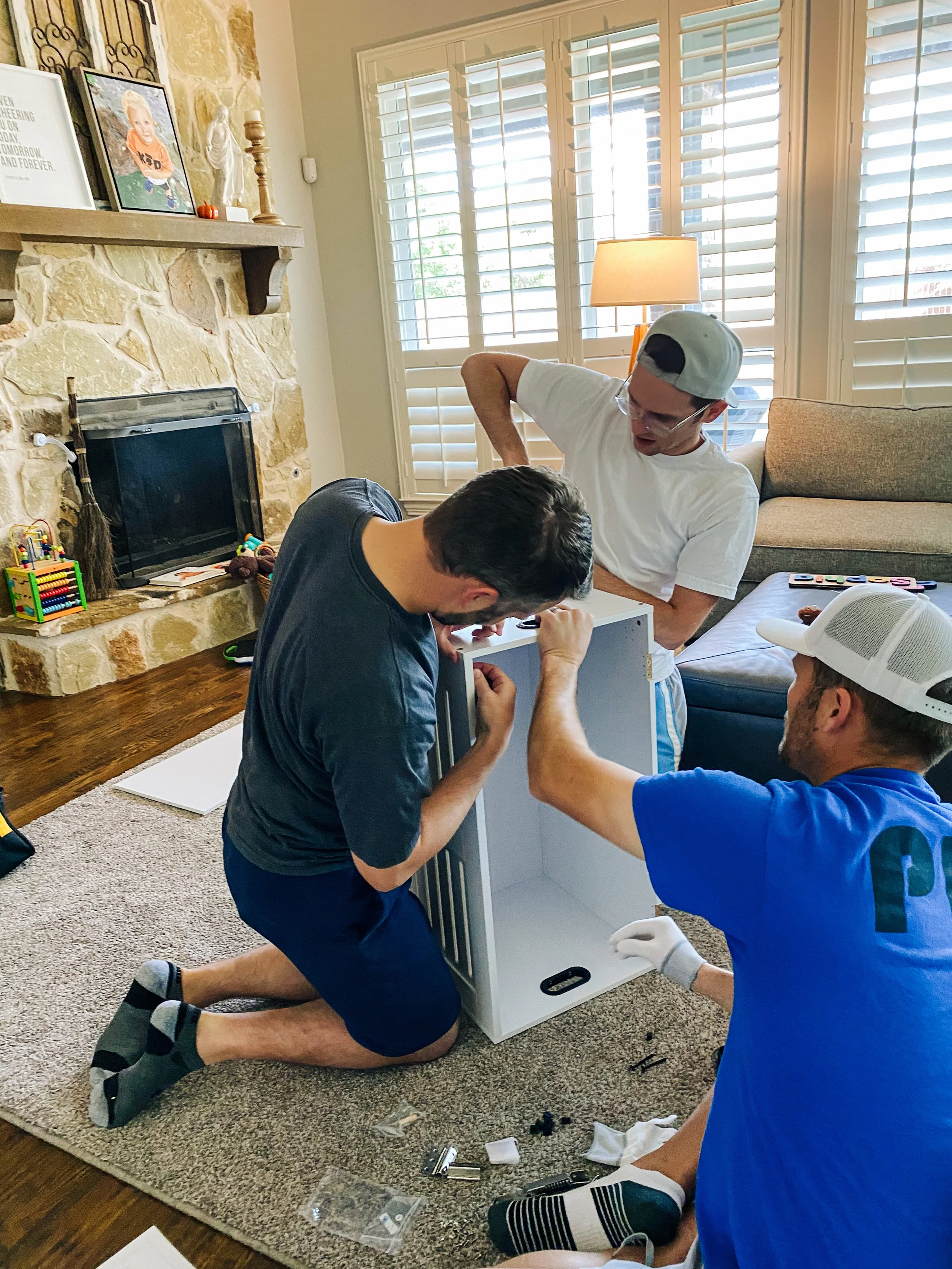 Three men assembling a small white cabinet in a living room with a stone fireplace, a beige couch, and a window with white shutters.