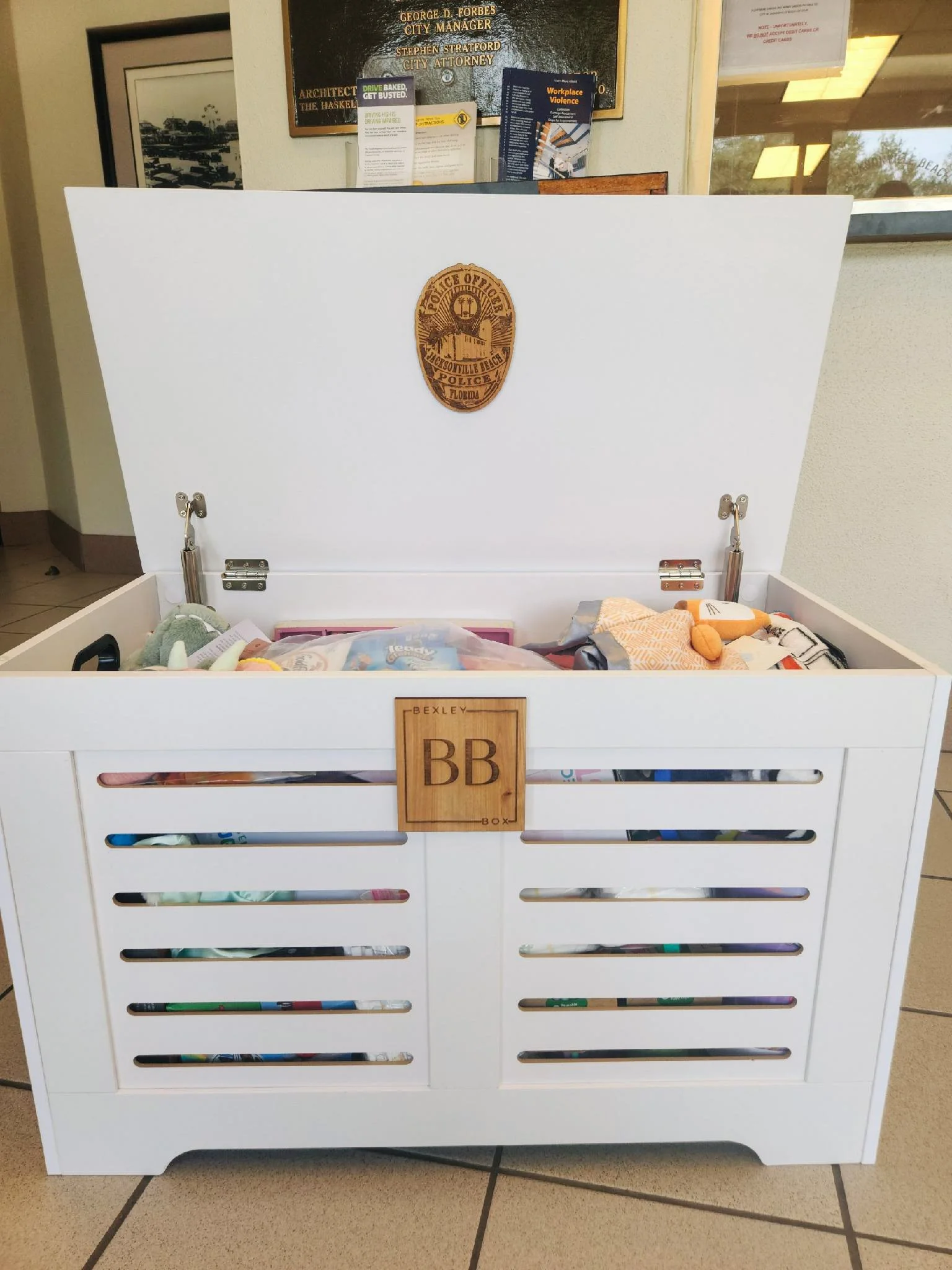 White toy storage chest with a police badge decal on the inside of the lid and a wooden plaque with castle and 'BB' initials on the front. The chest is open, showing stuffed toys inside. The background includes framed documents and posters on the wall.