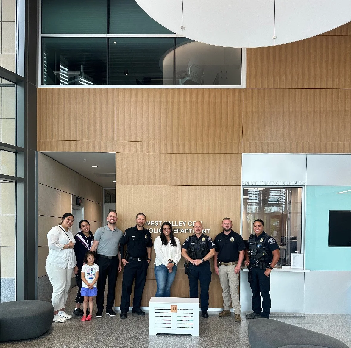 Group of seven people, including children, standing inside a police department lobby in front of a sign that says 'West Valley City Police Department'. Four of the adults are in police uniforms, and the others are in casual attire. The group is smili