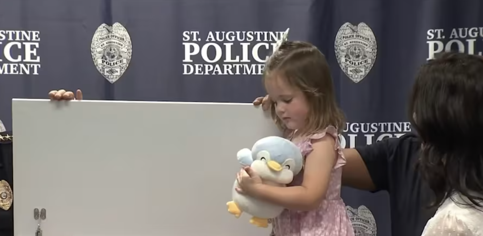 A young girl holding a plush penguin toy from a Bexley Box during a police event at the St. Augustine Police Department. An adult is partially visible alongside her. The backdrop features police department logos and insignias.