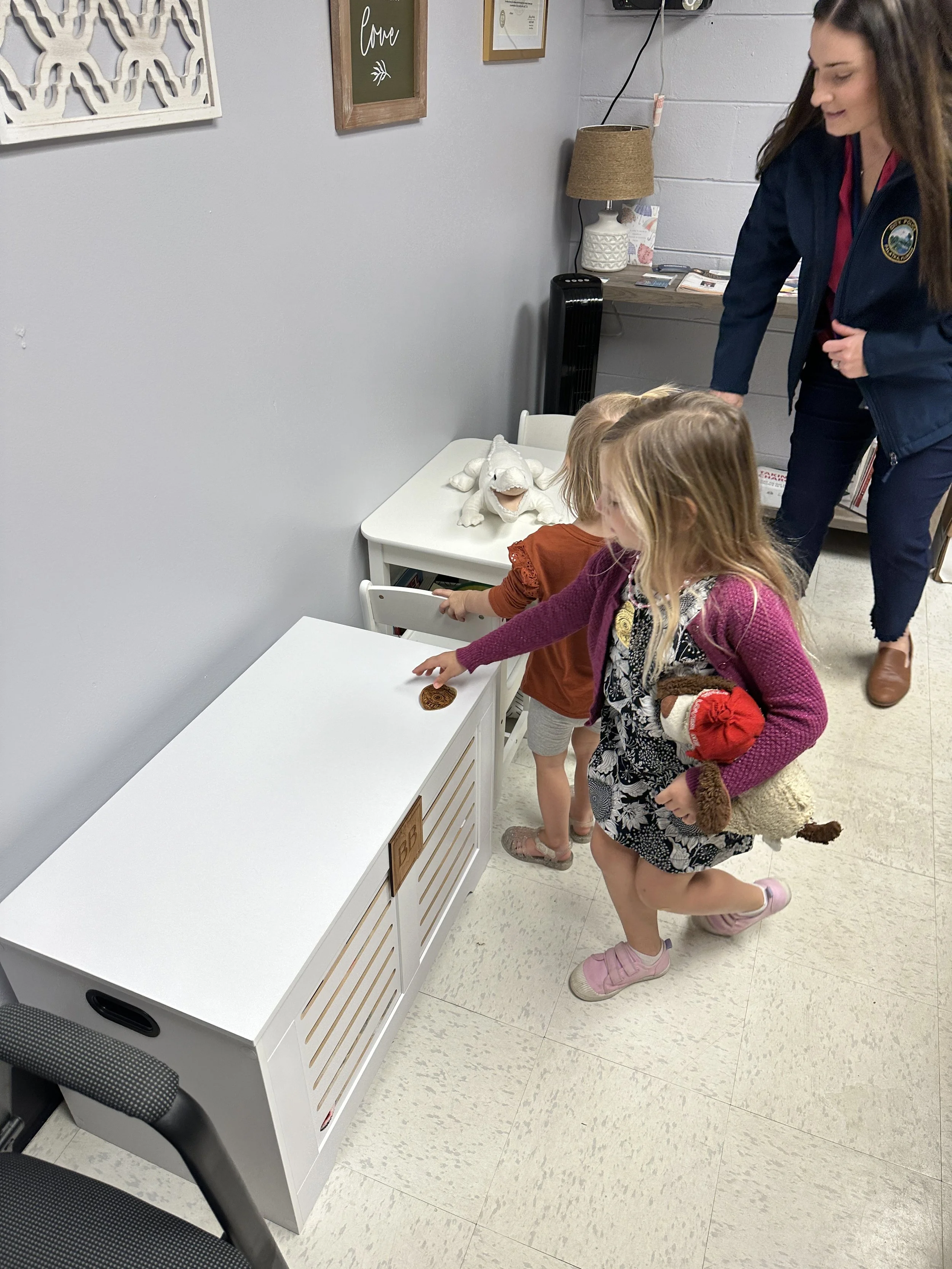 Young girl reaching for a cookie on a white storage bench, with two small children and an adult in a room decorated with framed art and stuffed animals.