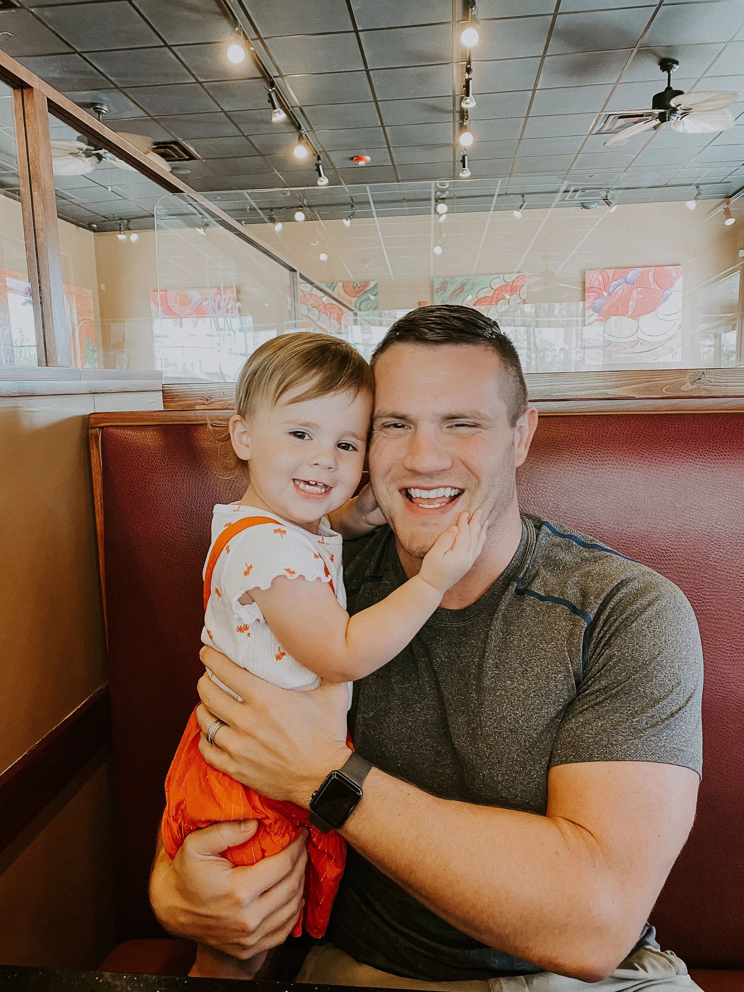 A smiling young child and an adult man are sitting in a restaurant booth, with the child touching the man's face. The child has short light brown hair and is wearing a white shirt with orange accents, while the man has short dark hair and is wearing a gray athletic shirt and a black smartwatch. The background shows part of the restaurant interior with colorful artwork on the walls and ceiling fans.