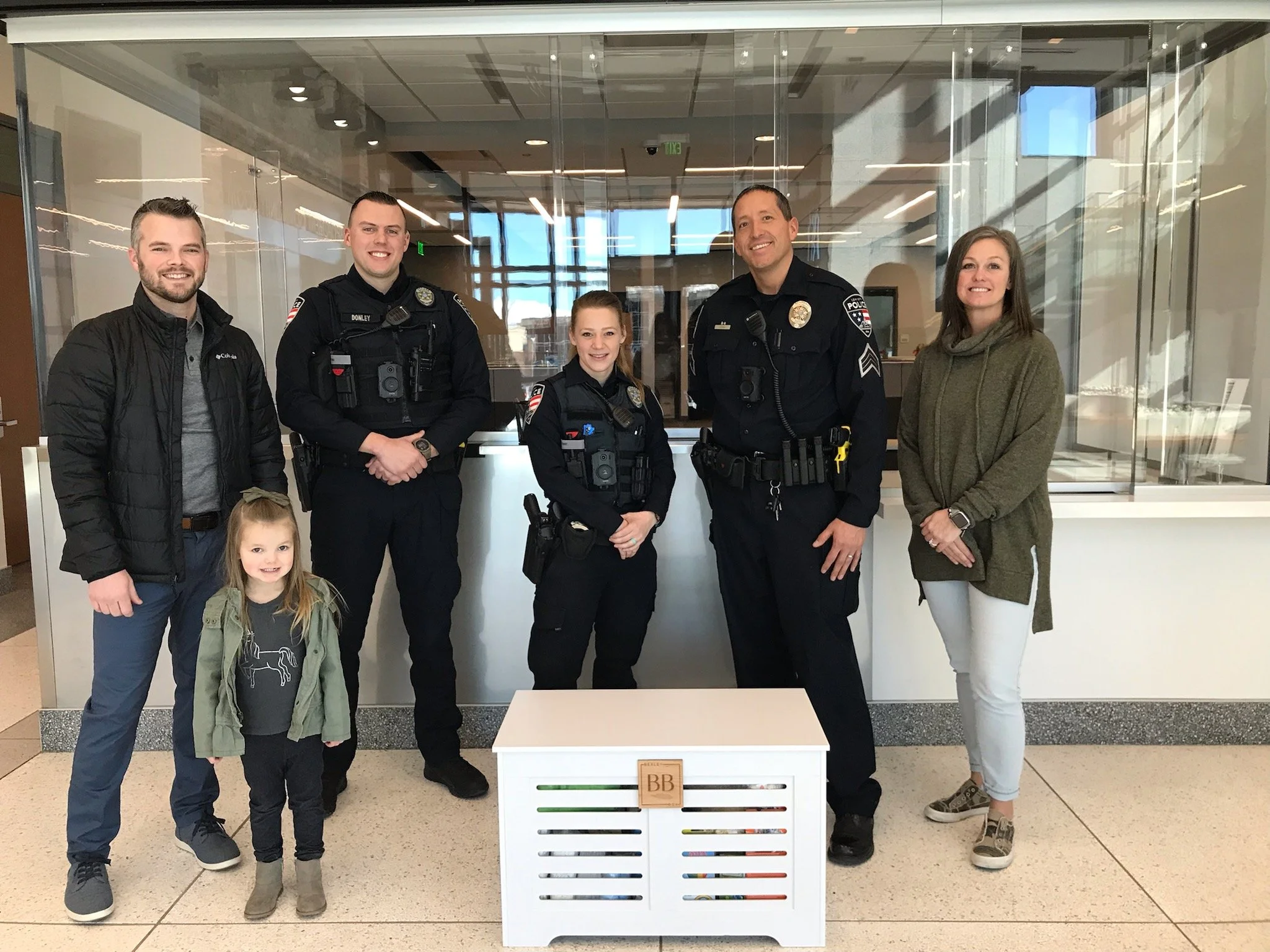 A group of six people, including two police officers and four civilians, standing inside a modern building with glass walls. Two children are in the front, and two women are on the sides. They are smiling and posing for the photo.