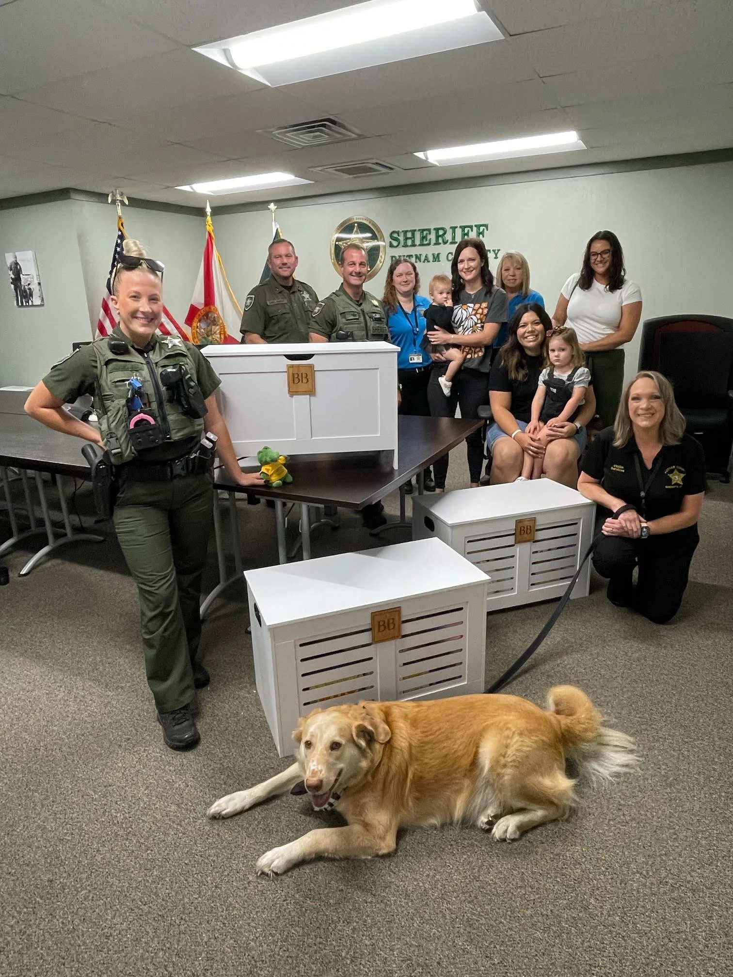 Group of police officers, sheriff staff, and family members posing in an office with a service dog lying on the carpet in front. The office has American and Florida flags behind the group and a sign that reads 'SHERIFF PASCO COUNTY.'