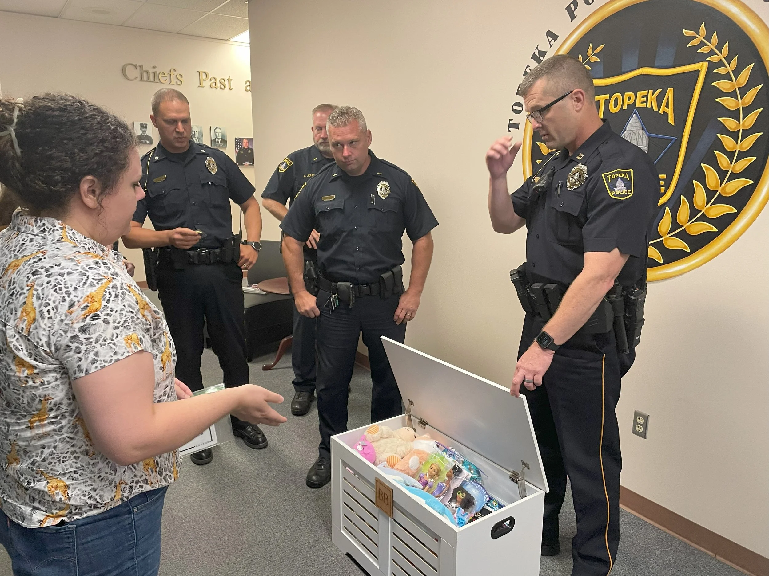 A woman with dark curly hair wearing a patterned shirt receives a donation of toys from a group of five police officers standing in front of a police emblem wall.