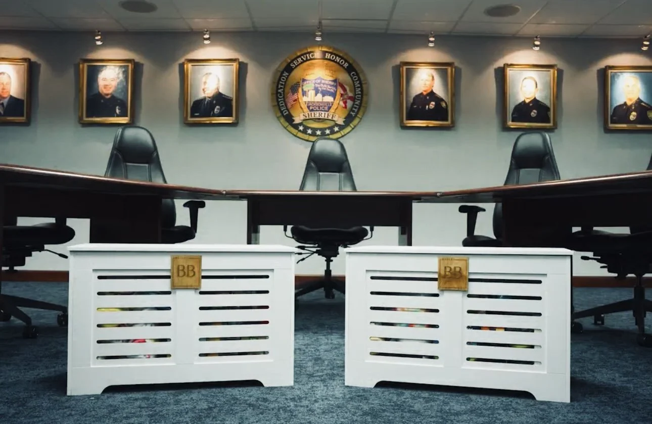 Empty sheriff's office conference room with a large central table, black chairs, framed police portraits on the wall, and a sheriff's badge emblem displayed in the center.