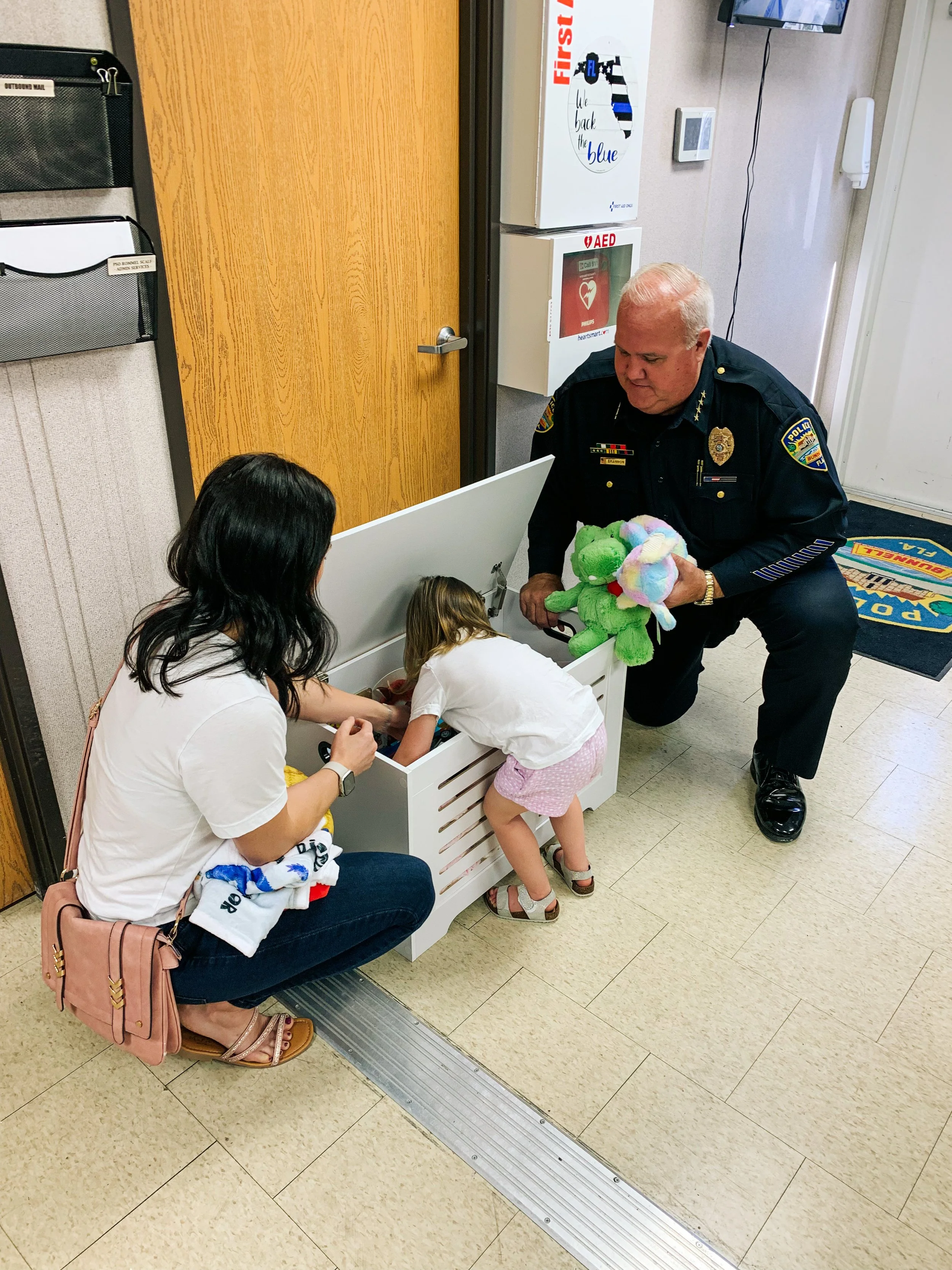 A police officer kneeling on the floor hands a stuffed toy to a young girl reaching into a white toy chest, while a woman sitting on the floor watches. The setting appears to be a school or community center.