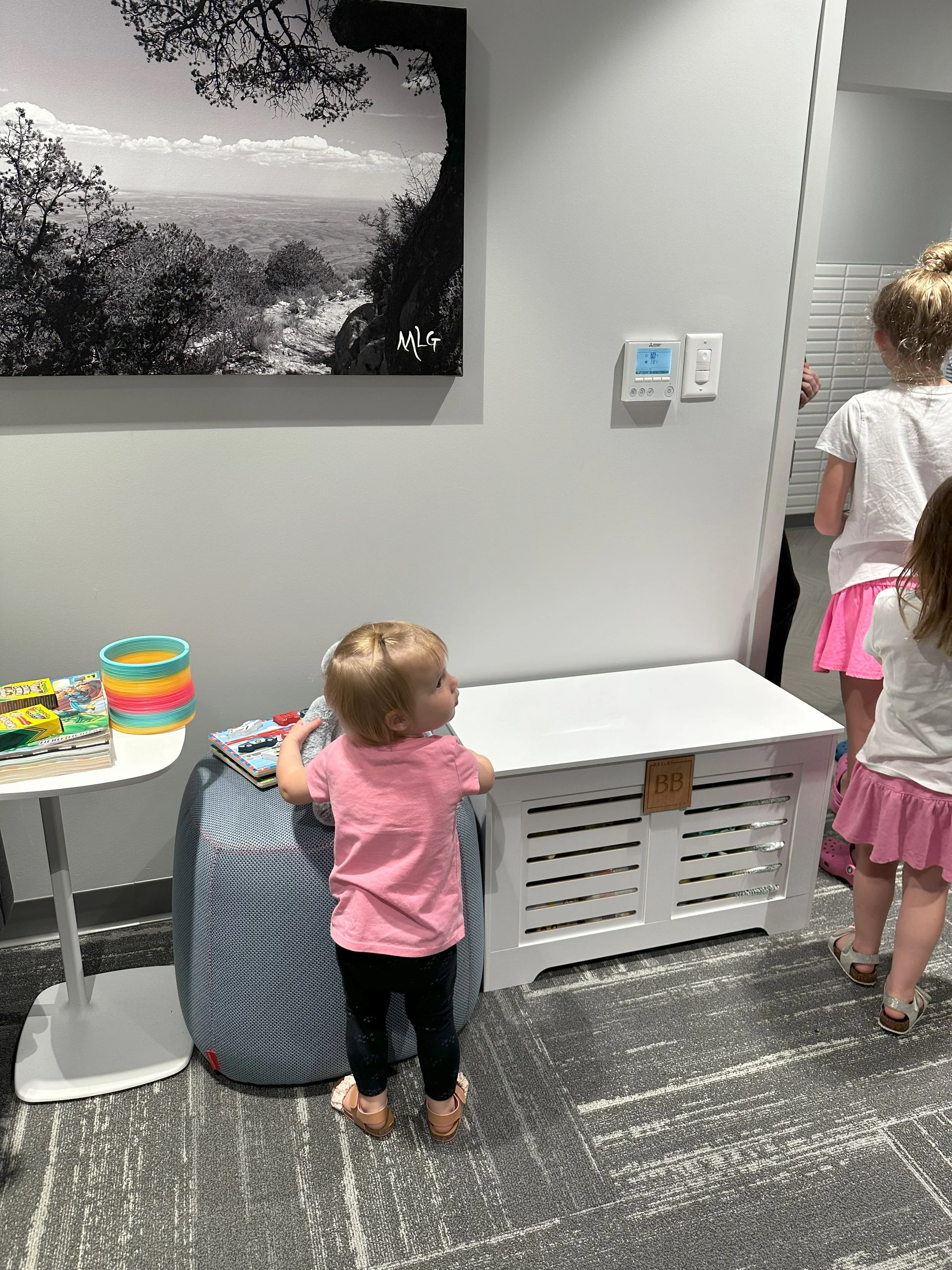Three young girls in pink and white are standing and looking at a white radiator cover in a room with gray walls and dark striped carpet; one girl with a pink shirt and black pants is leaning on a gray cushion against the wall, while two other girls in white shirts and pink skirts are near the radiator and hallway. To the left, a small white table holds colorful plastic cups and some books.