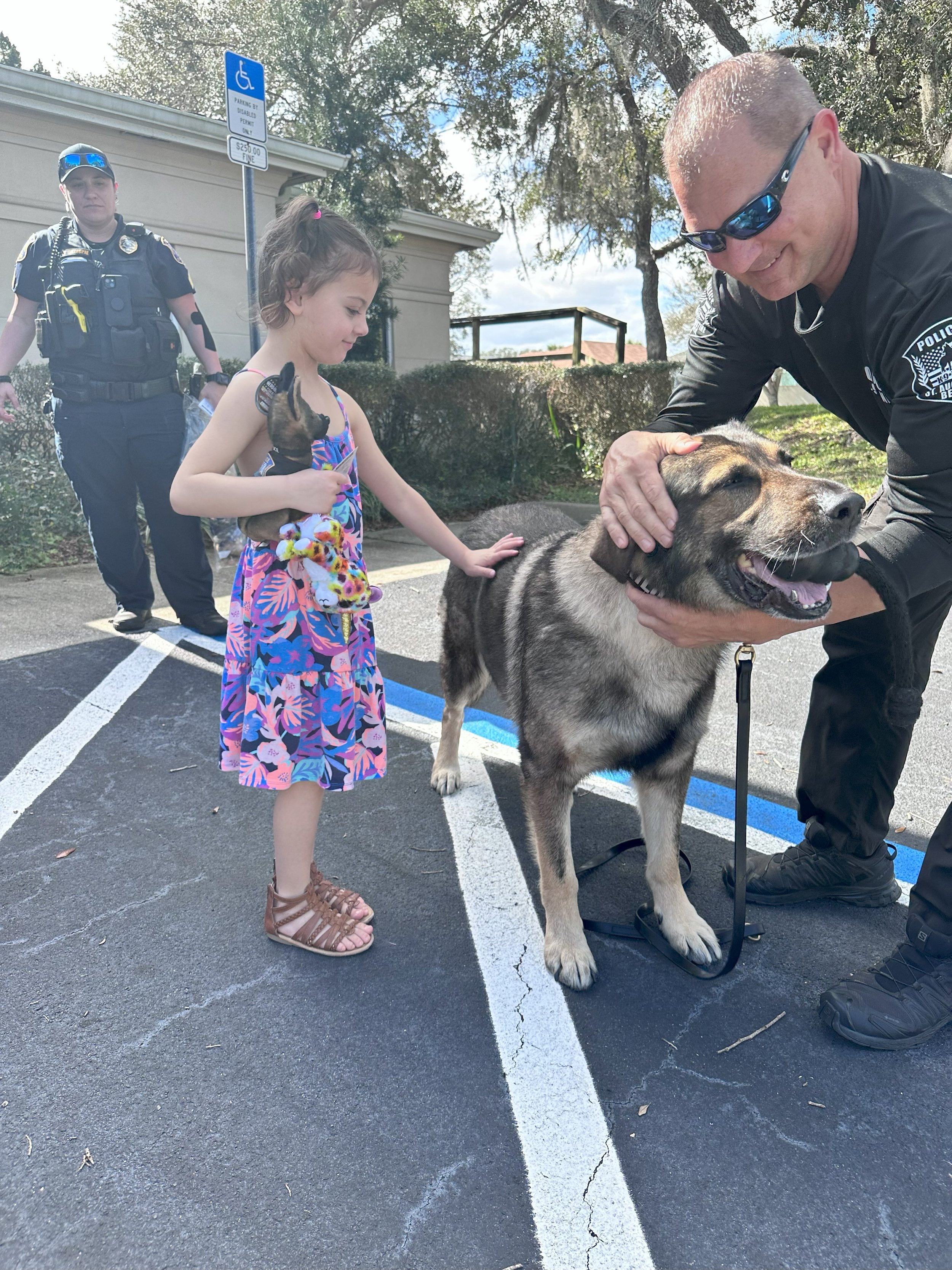A young girl in a colorful dress petting a police dog while a police officer bends down to hold the dog's head. A police officer stands in the background near a parking space marked for handicapped with a blue sign.