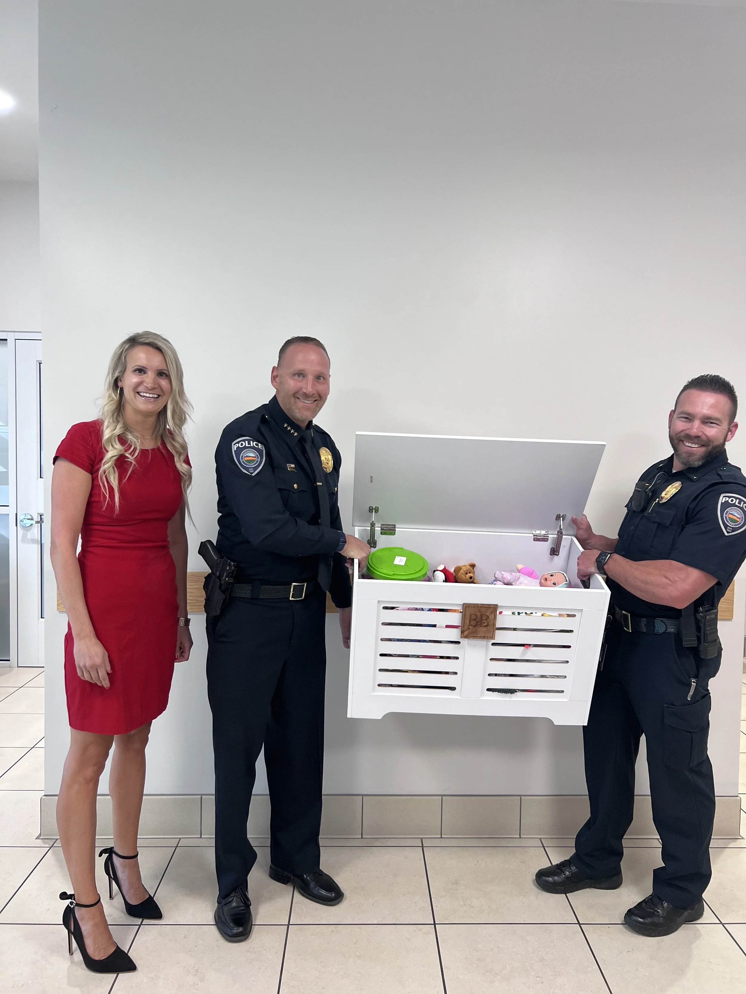 Three people, including two police officers in uniform and a woman in a red dress, standing next to a white toy chest filled with dolls and toys in an indoor setting.