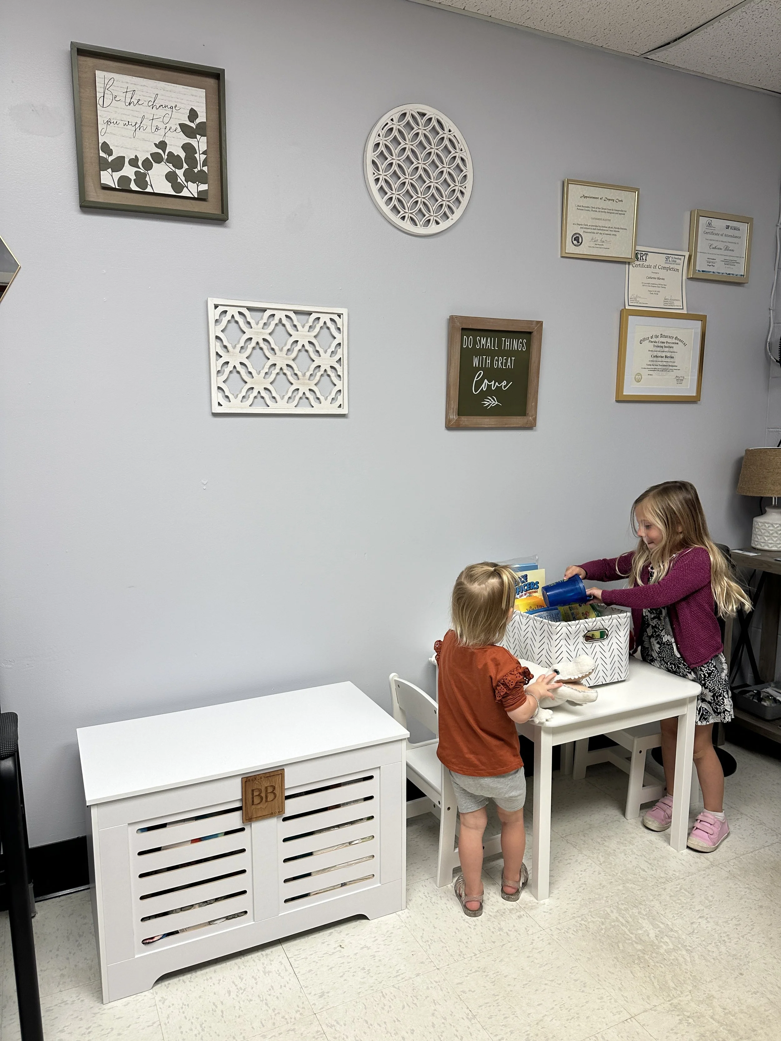 Two young girls are playing with toys and a box of books at a white table in a room with framed decorations and certificates on the wall.