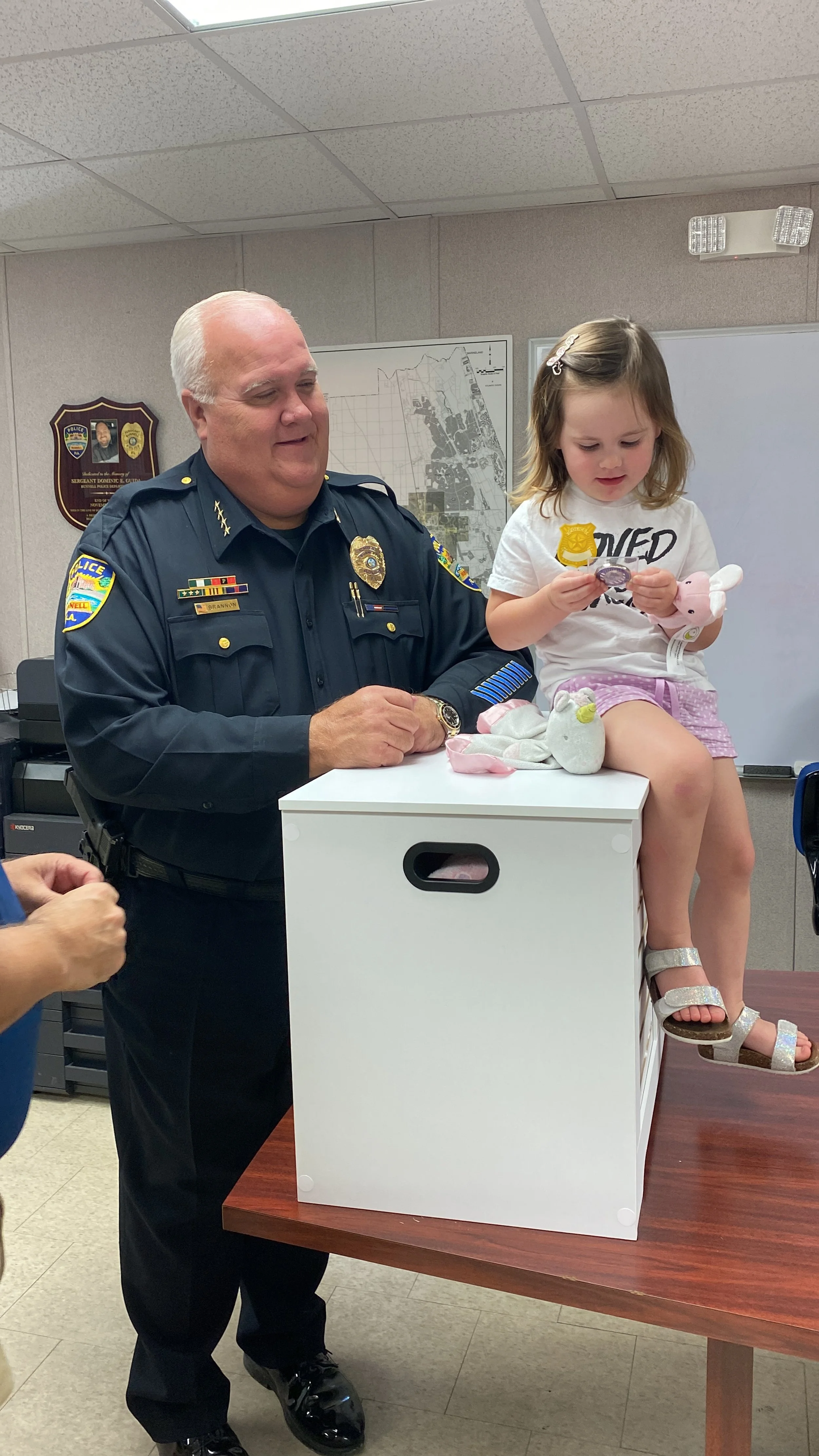 A police officer standing next to a young girl sitting on a white storage cube, holding a pig stuffed animal and some coins, during a community event in an indoor setting.