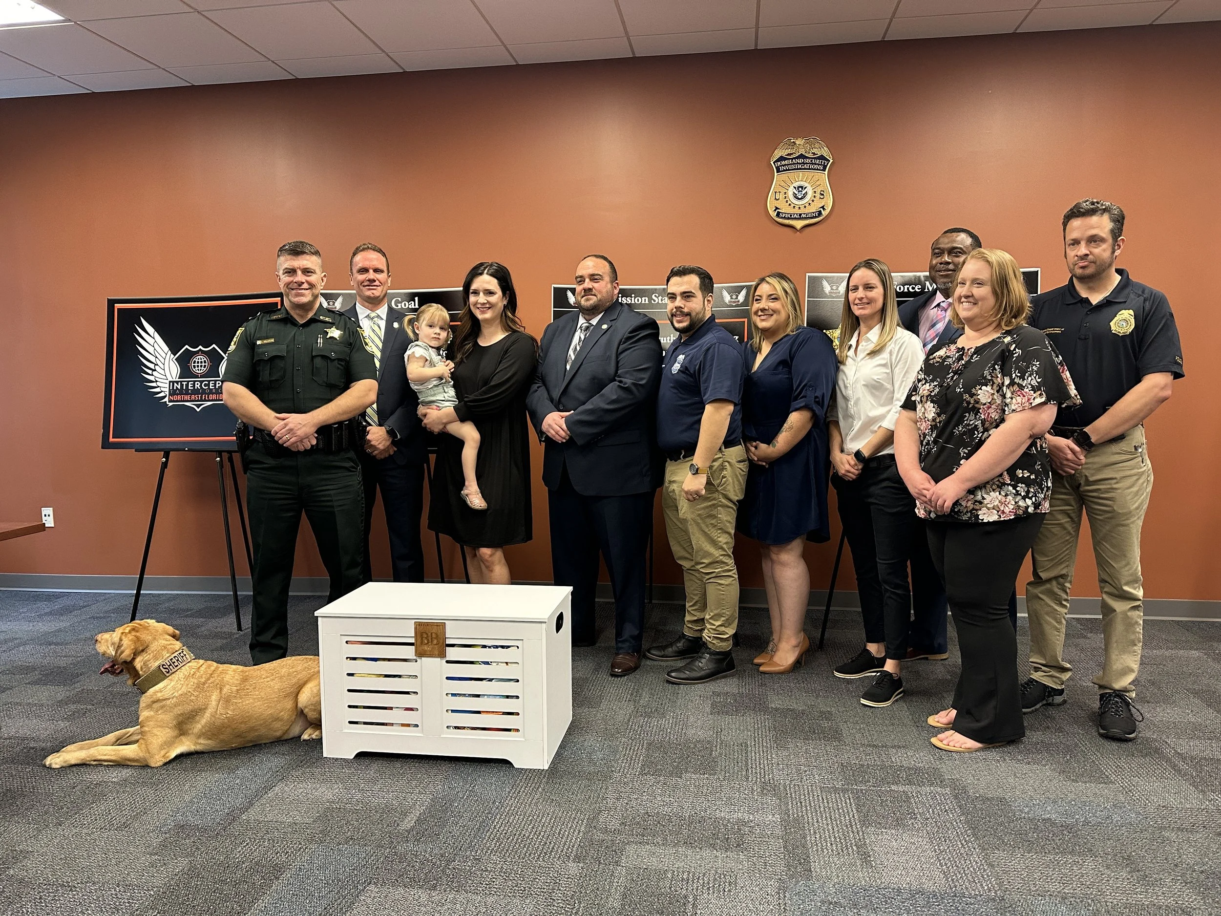 Group of people and a dog in a room with an orange wall and police logos, some standing and one man in a sheriff's uniform, accompanied by signs and a display.