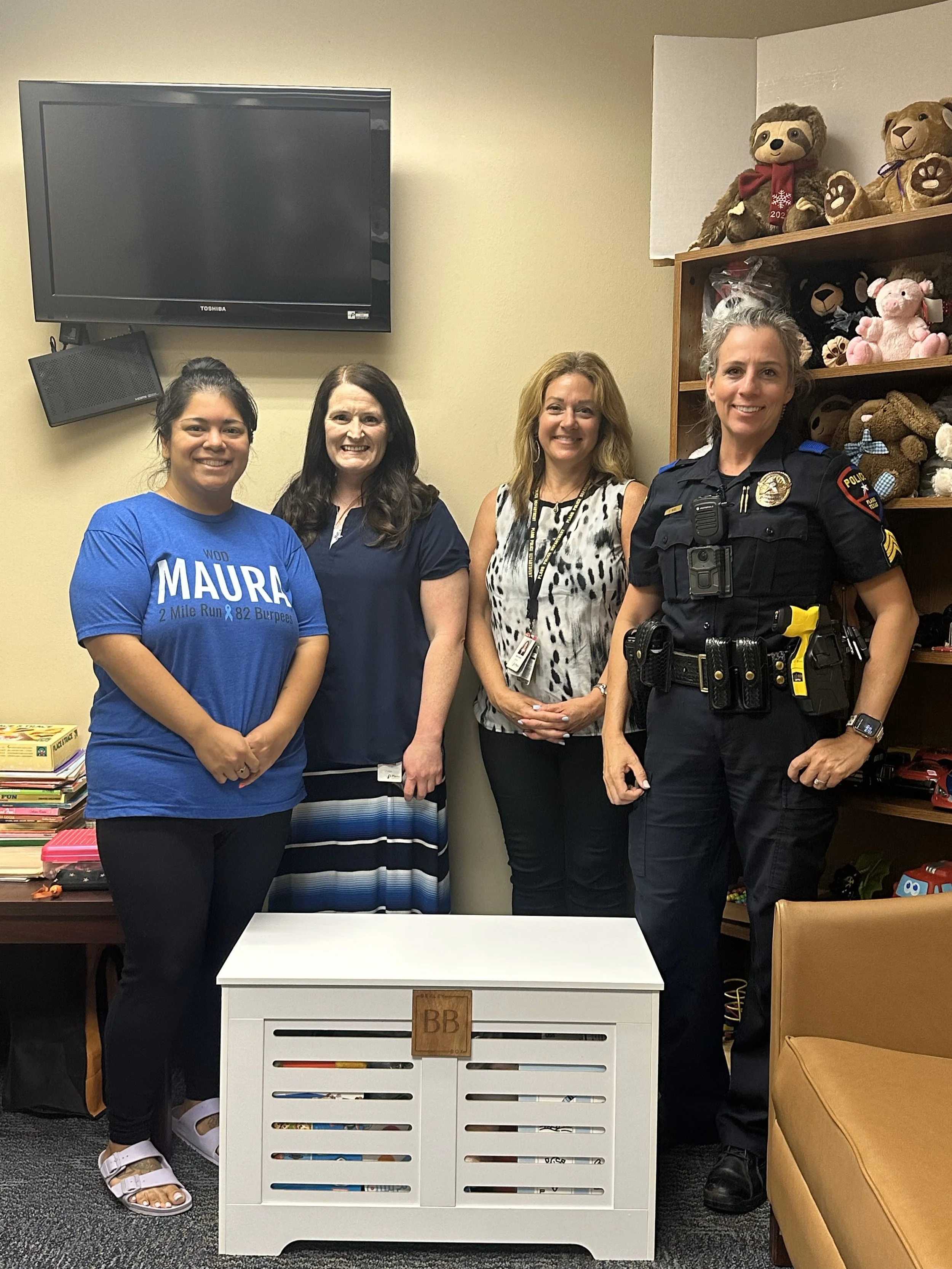 Four women standing in an office or room with toys on a shelf behind and a television mounted on the wall. One woman is in police uniform, and the others are dressed in casual or business casual attire. There is a small white wooden bench with the in