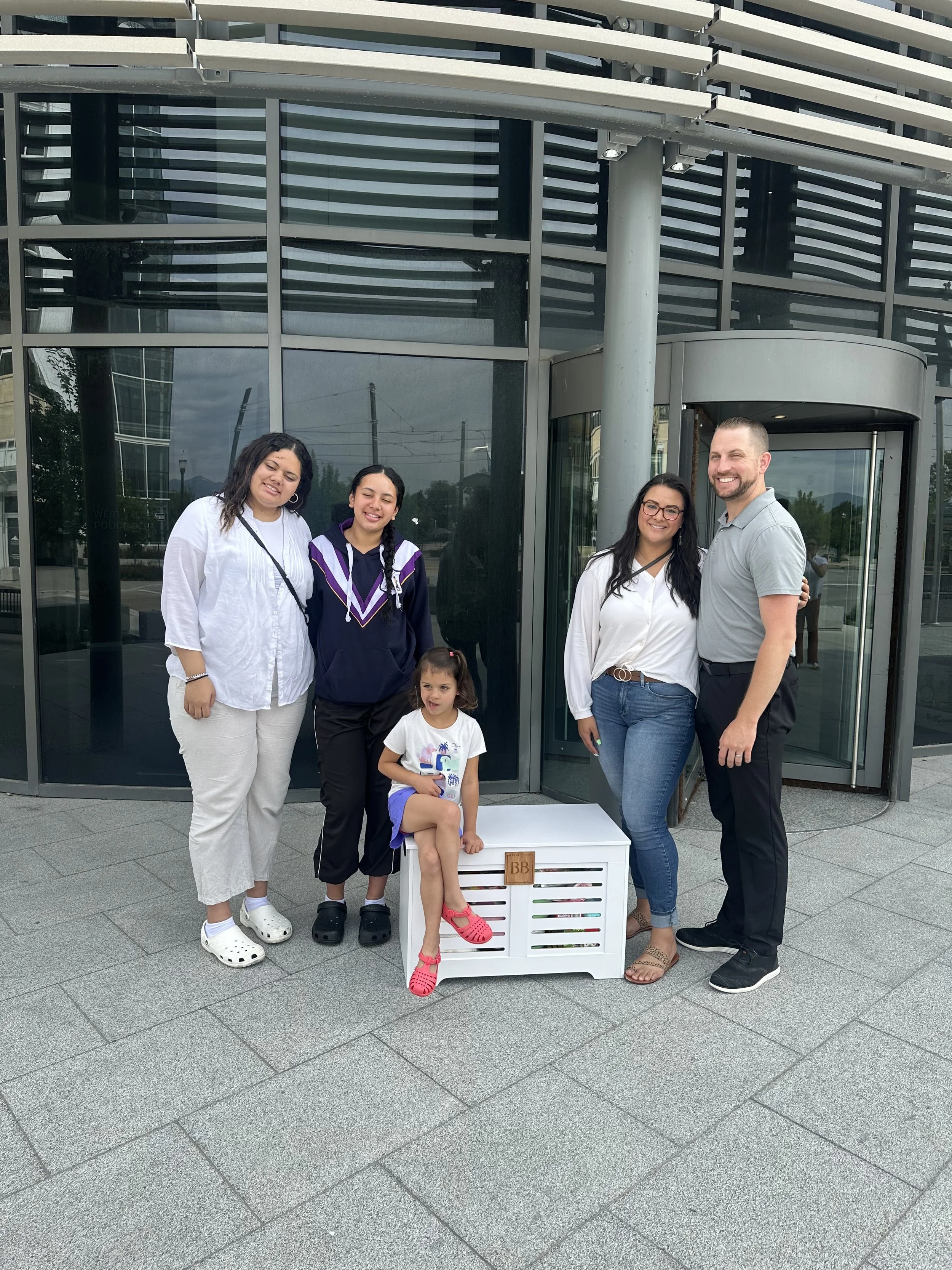 Group of five people, four women and one man, standing outdoors in front of a modern glass building, smiling for the camera. One young girl sits on a white crate labeled 'BB' with pink sandals.