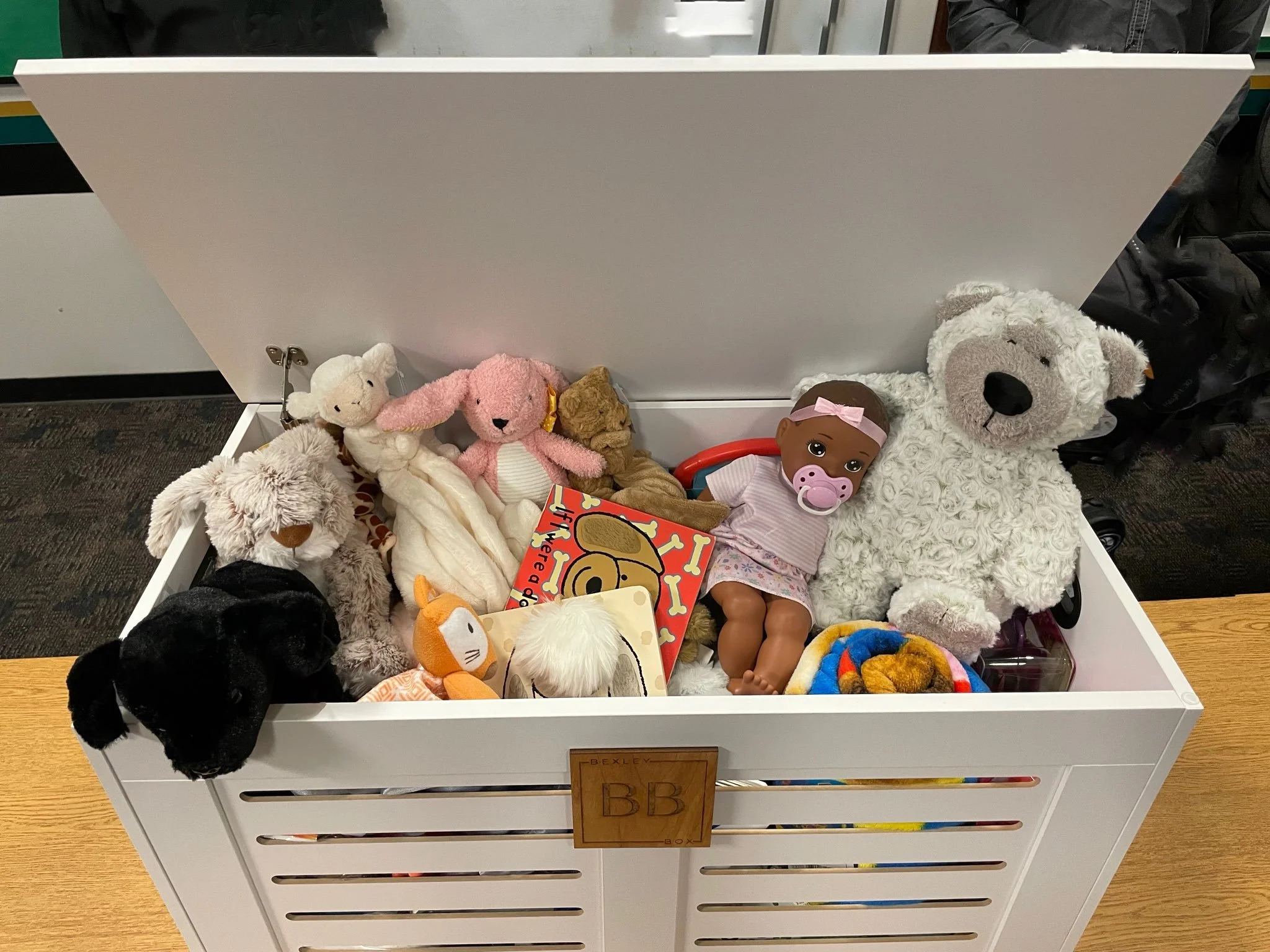 A white storage box filled with plush toys and a doll, with a wooden label that says "BB."