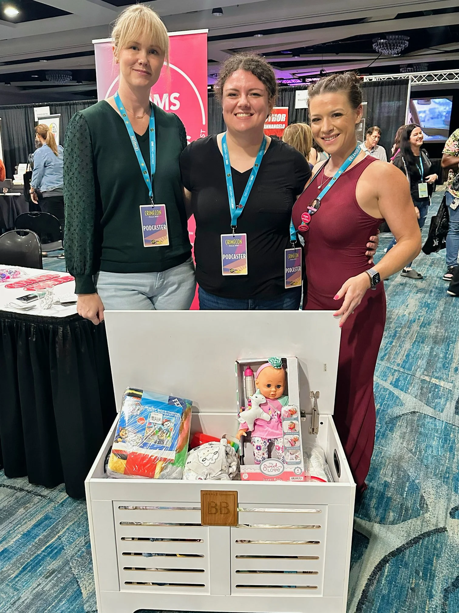 Three women at a convention standing behind a white crate filled with toys, including a doll and other items, all wearing conference badges.