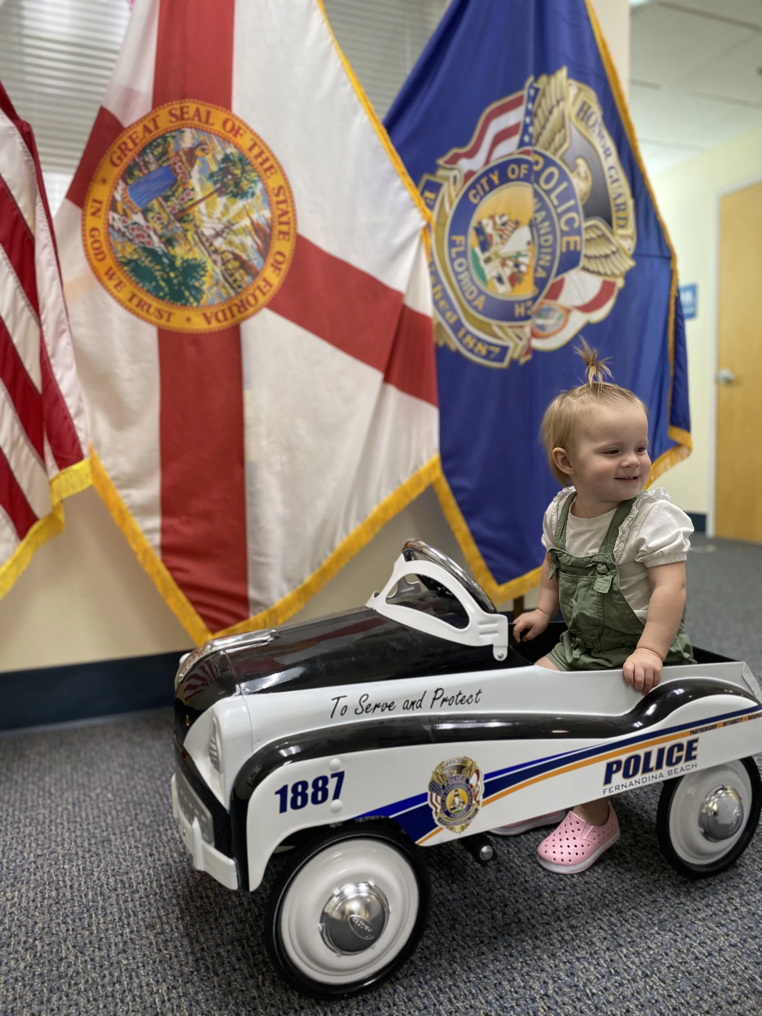 A young girl sitting in a small toy police car marked with 'To Serve and Protect' and 'POLICE FERANDINA BEACH,' with police flags in the background.