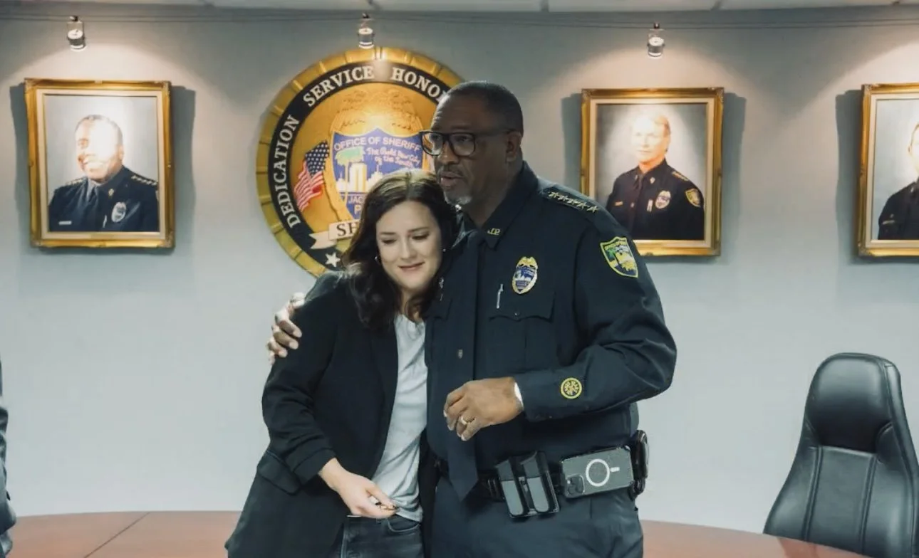 A woman hugging a police officer in a conference room with portraits of officers on the wall behind them and a sheriff's badge emblem.