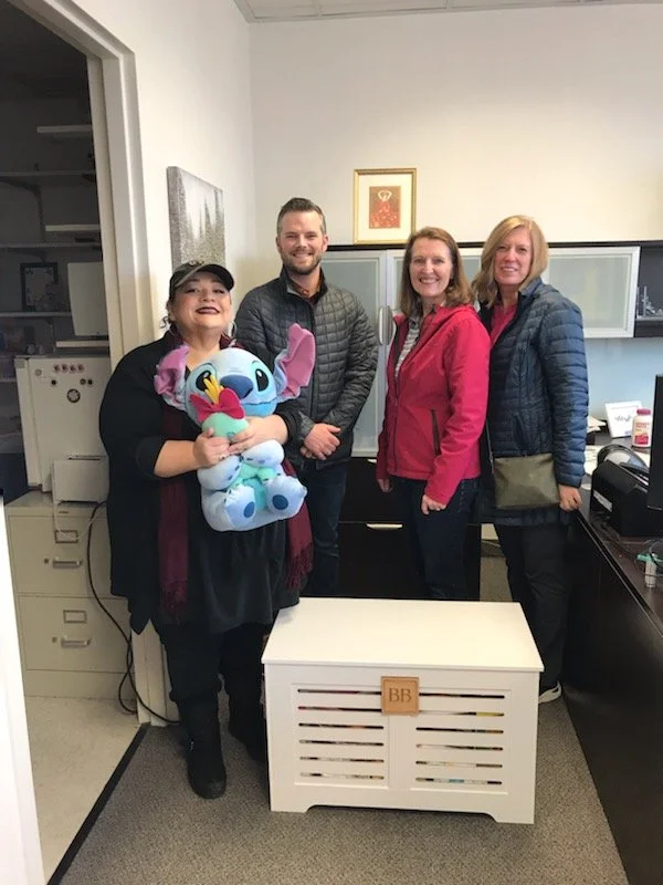 Four women standing in an office, with one holding a Stitch plush toy. They are smiling and posing for the photo.