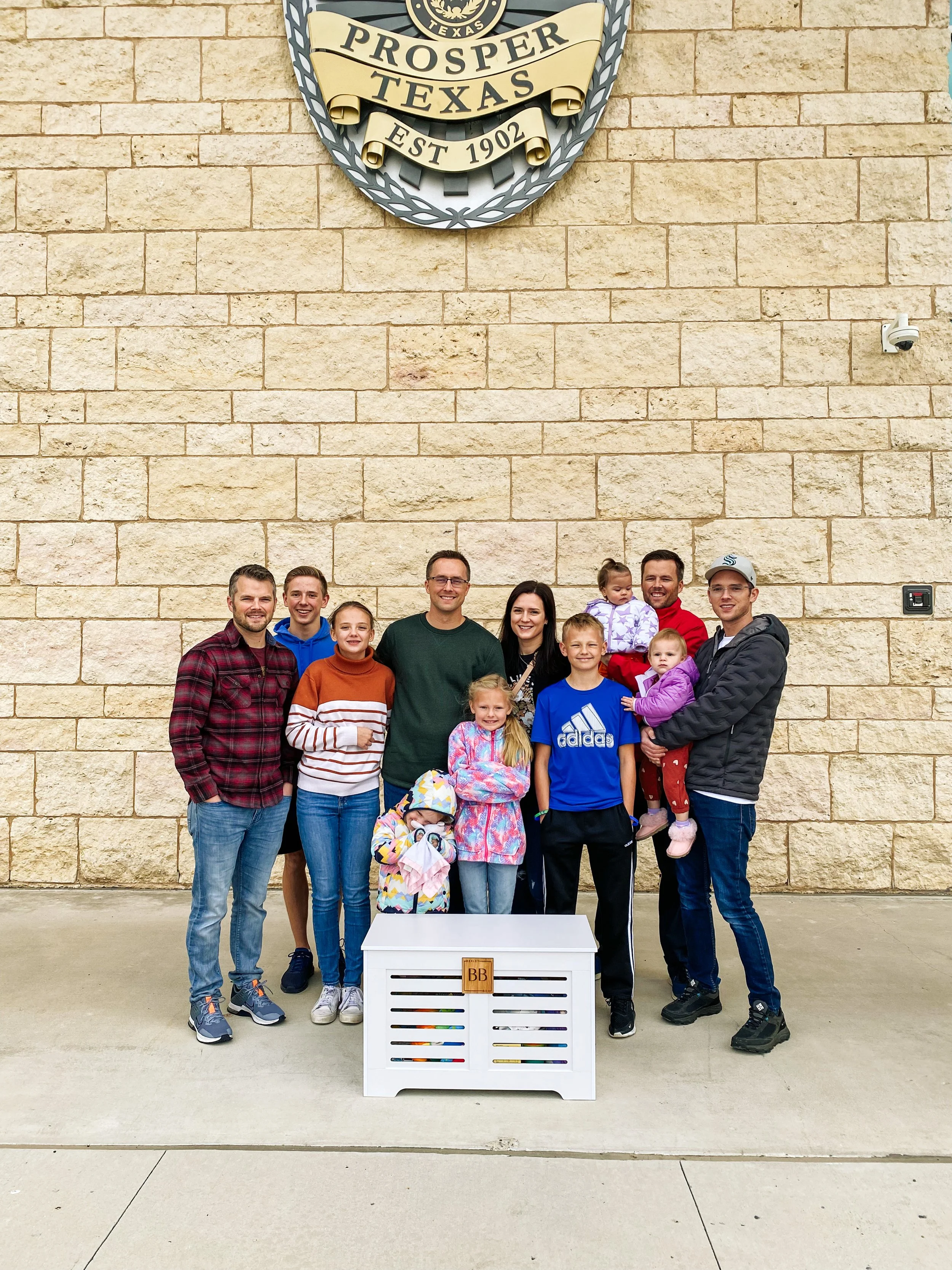 Group of people, including children, standing in front of a brick wall with a Texas-themed emblem that reads 'Prosper Texas' and 'EST 1902.'