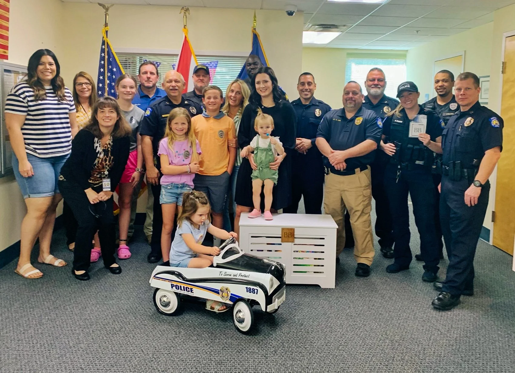 A group of children and police officers posing together indoors, with flags in the background, some children sitting or standing on a white crate and a toy police car in front.