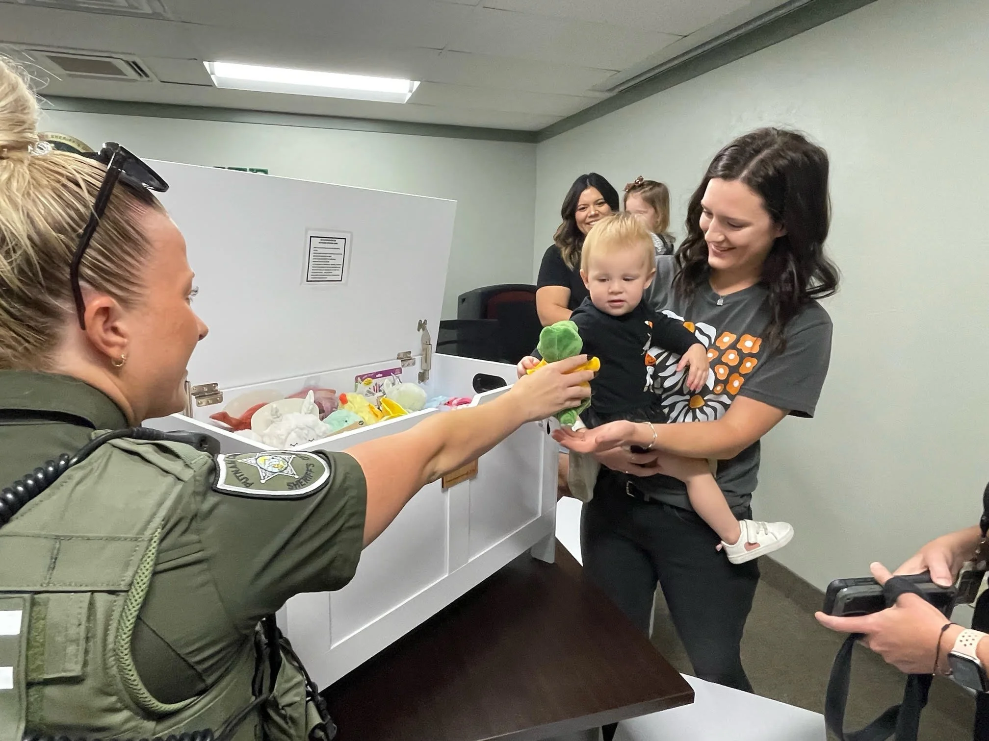 A police officer handing a stuffed animal to a woman holding a young child in a room, with other women and children in the background.
