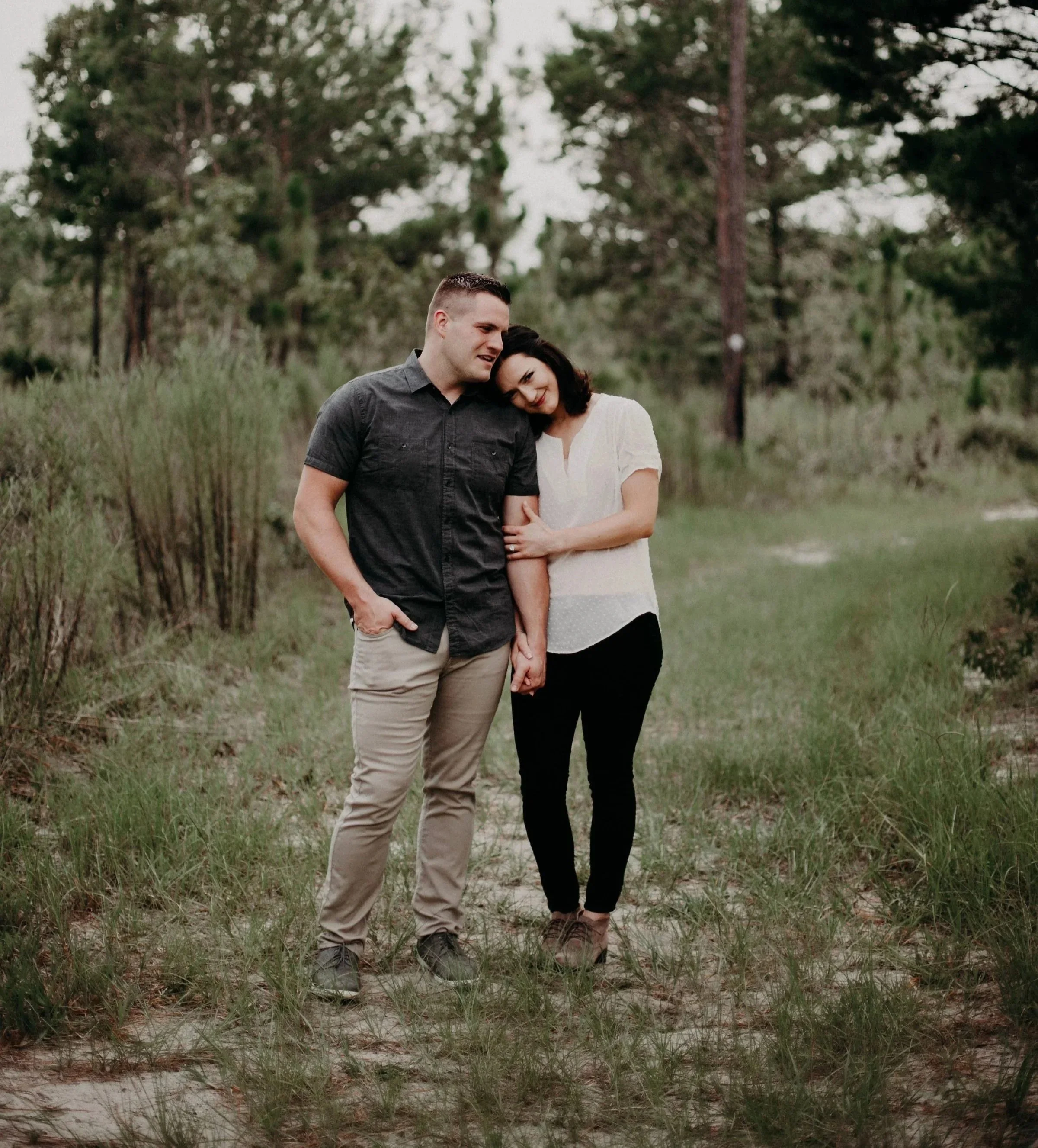 A man and woman standing closely together on a grassy path in a natural outdoor setting with trees and bushes in the background. The woman is resting her head on the man's shoulder, and they are holding hands.