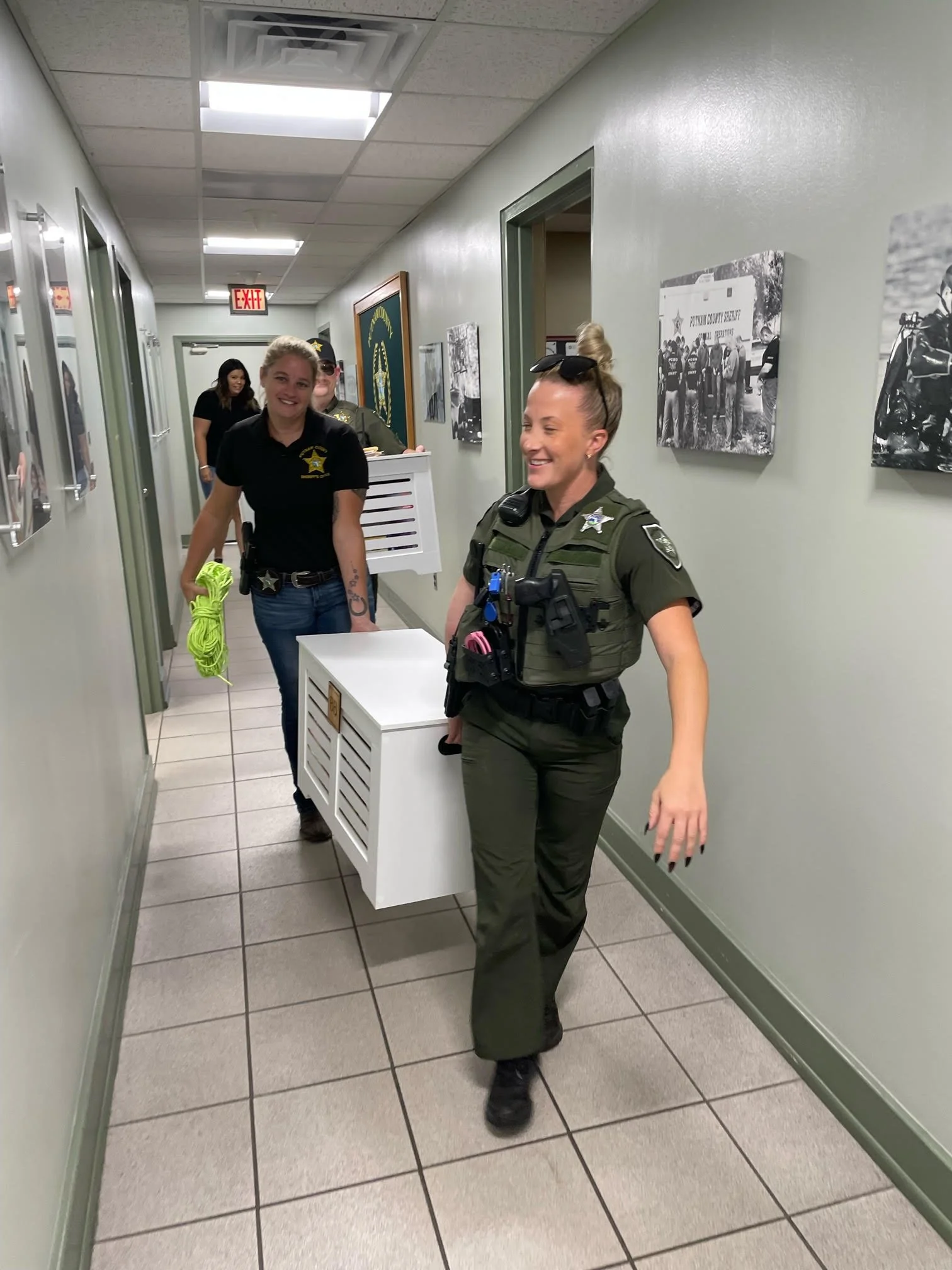 Several female police officers walking down a hallway with framed photos on the walls, smiling and carrying equipment, in a police station.
