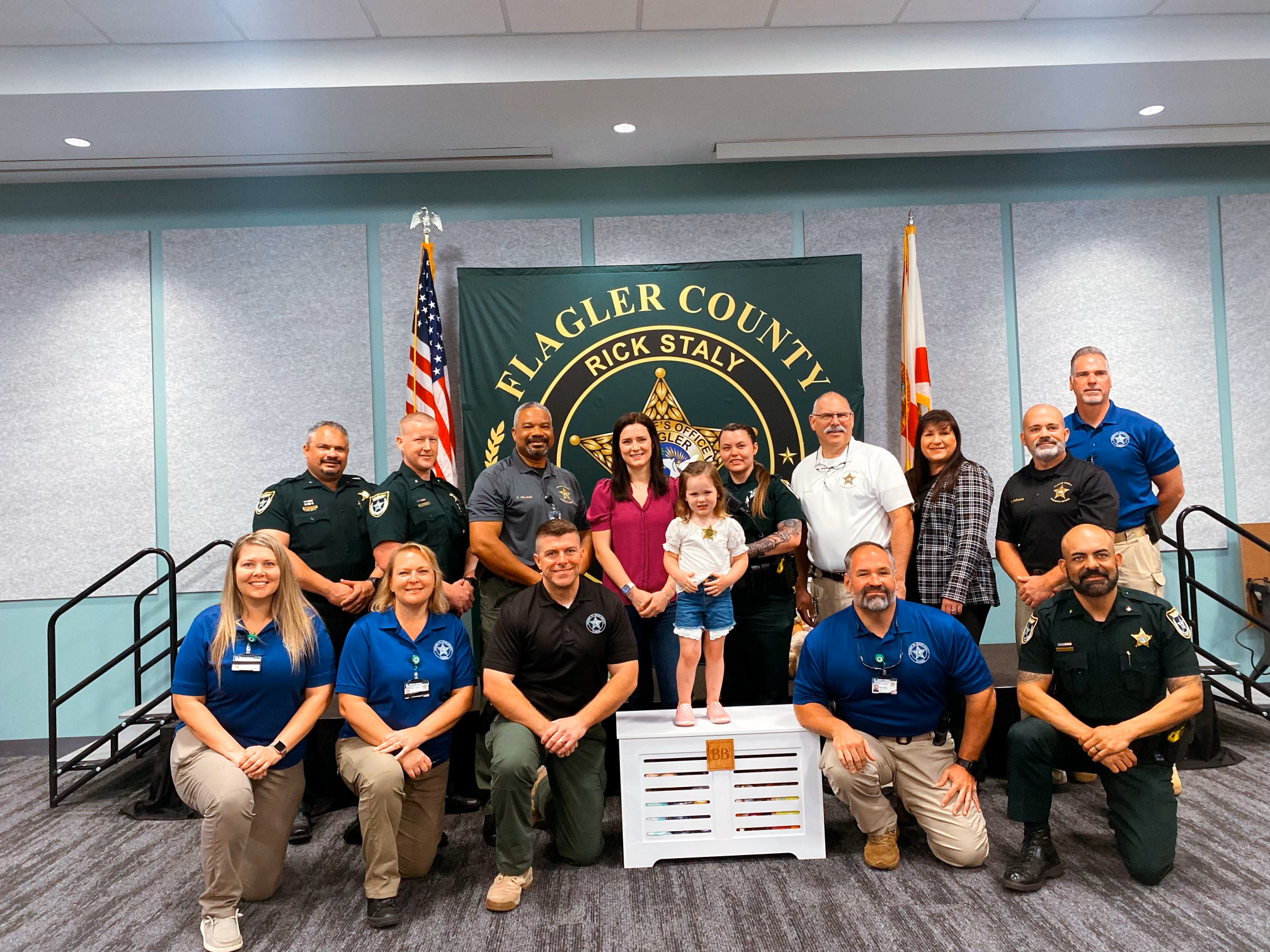 Group photo of law enforcement officers and community members in front of a Flagler County banner, with American flags, inside a conference room.
