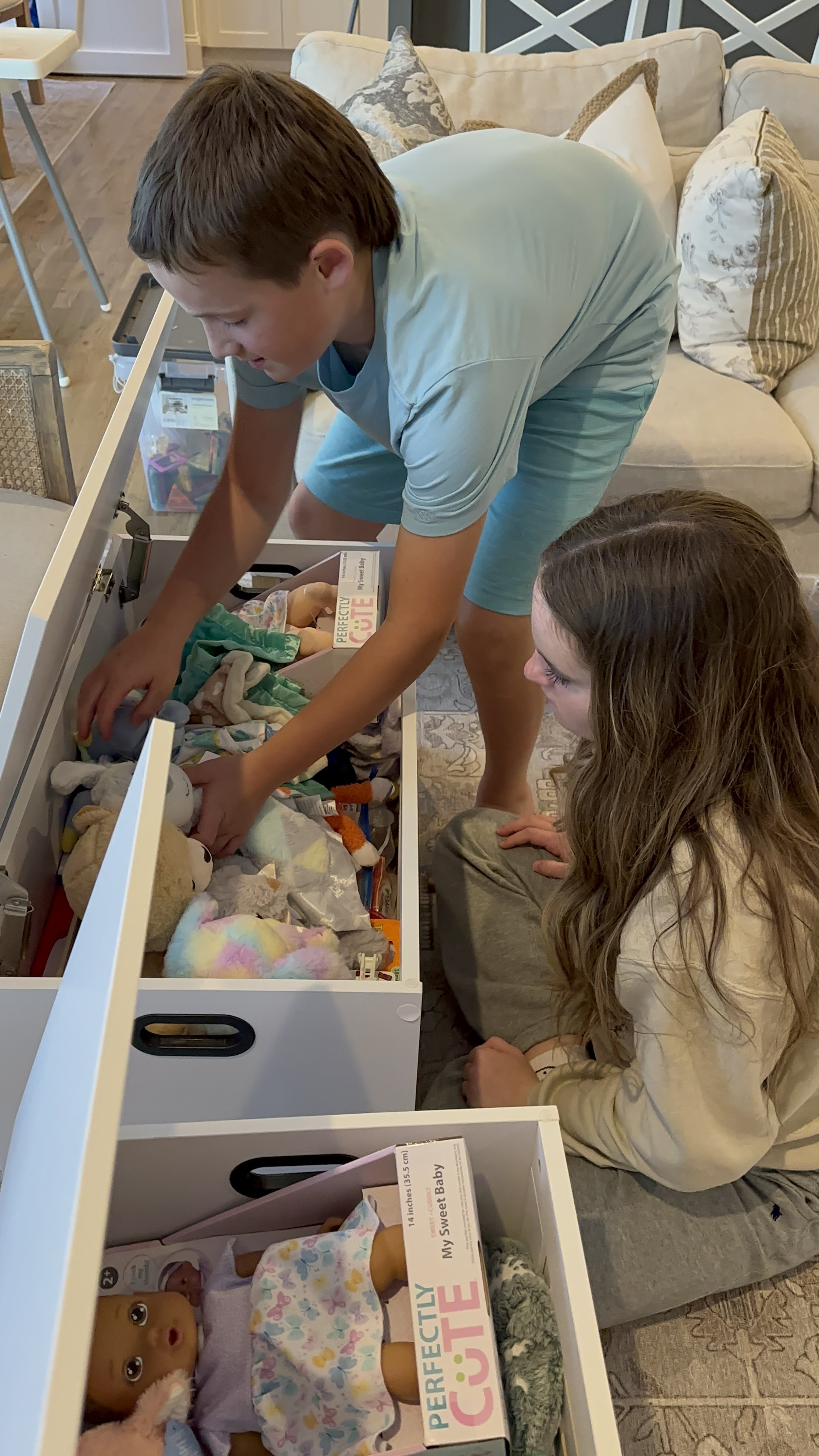 A boy and a girl looking into a toy chest filled with stuffed animals and toys. The boy is reaching into the chest, and the girl is sitting beside him on the floor. Both are indoors, in a living room with a beige sofa and patterned pillows in the bac