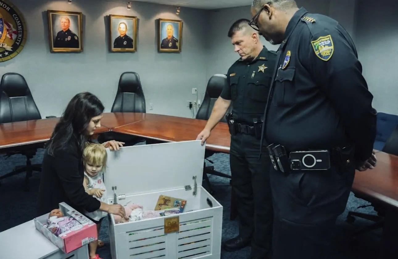 A woman and a young girl are looking at a white wooden box with children’s items inside, while two police officers stand nearby in a conference room with framed photographs on the wall.