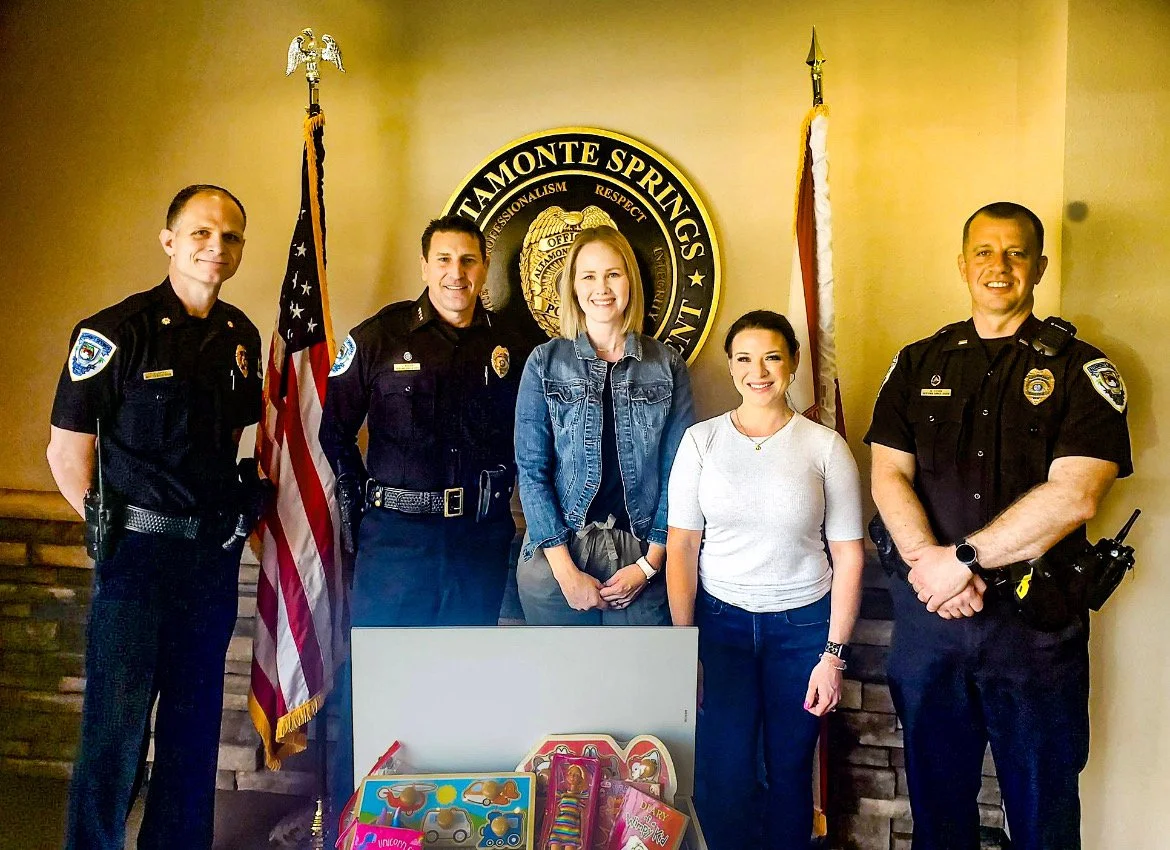 Four police officers and two women standing in front of a ceremony plaque and flags, with gift bags and toys on a table in front of them.