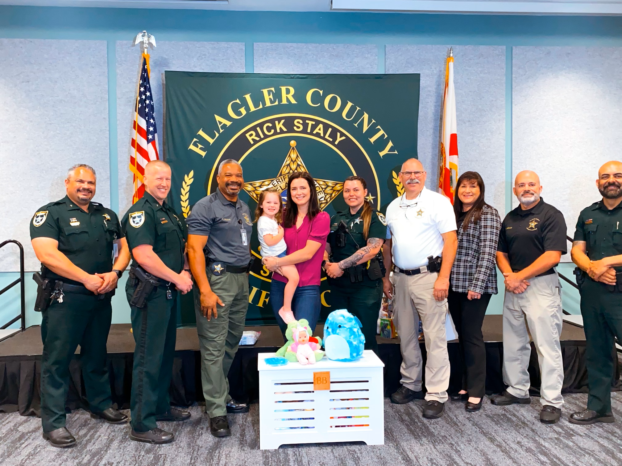 Group of sheriff's deputies, a woman and a young girl standing in front of a Flagler County sheriff's department backdrop, with American and state flags, at a ceremony or event.