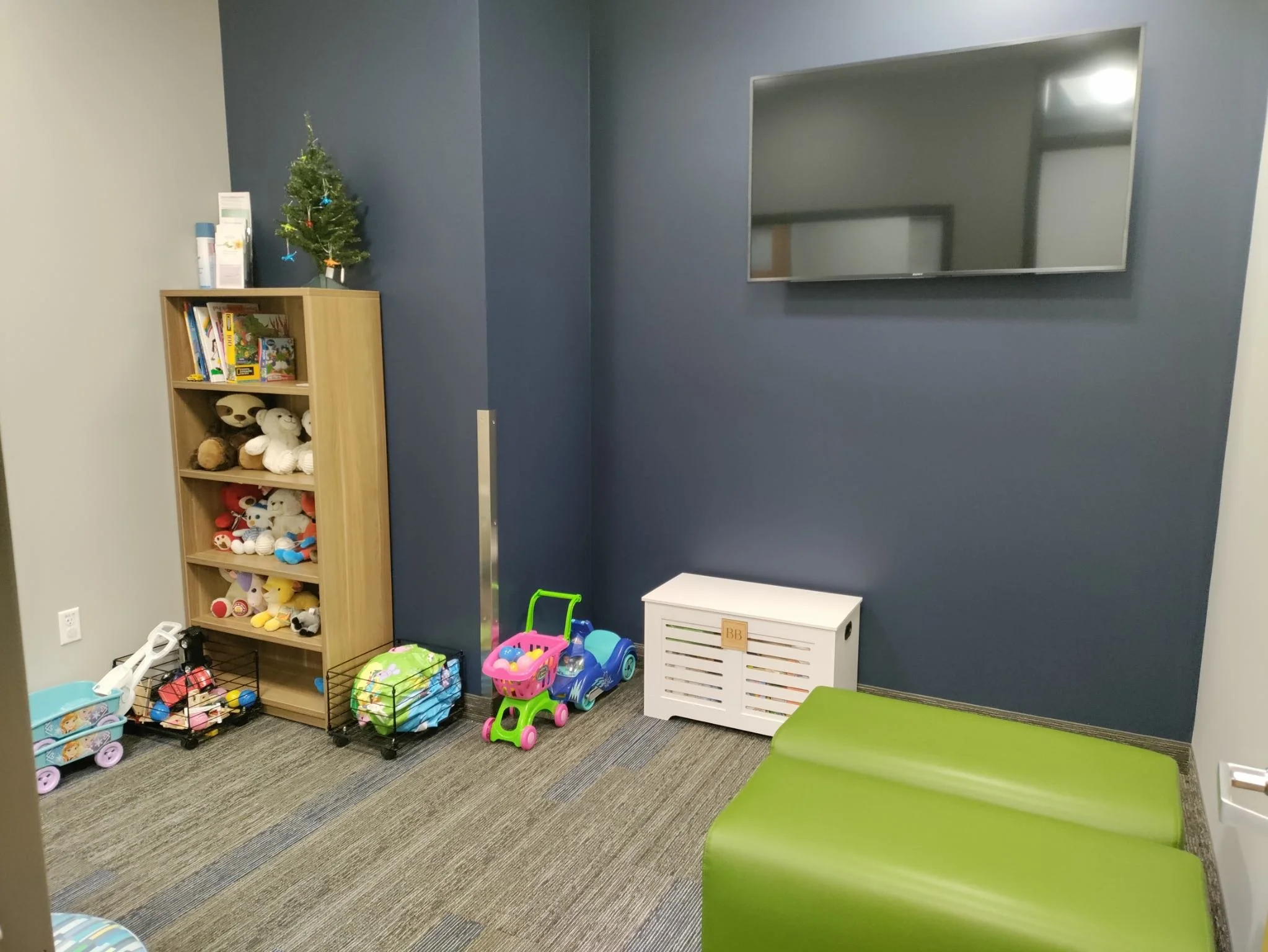 Children's playroom with a wooden bookshelf filled with toys and books, a tiny Christmas tree on top, colorful toy shopping carts, a small ride-on toy car, a white storage bench, and a green cushioned seat against a dark blue wall with a mounted flat