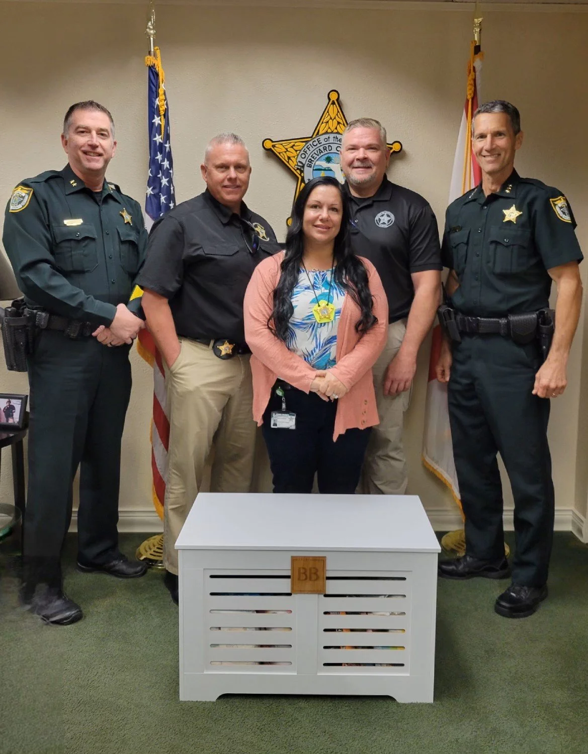 A group of five police officers and a woman standing in front of a police badge and flags, with a white bench in the foreground.