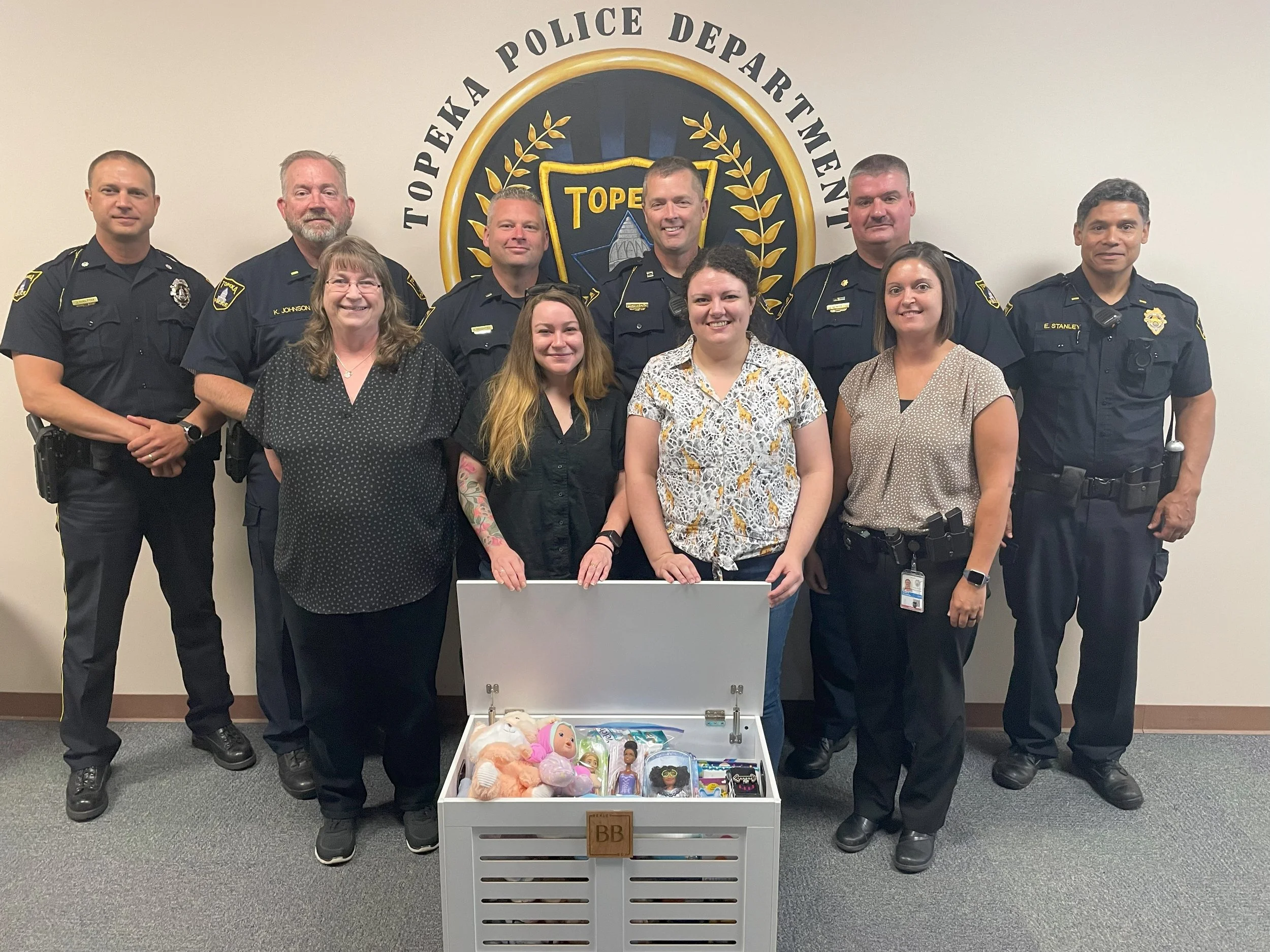 Group photo of ten police officers and staff members standing in front of the Toppen & Topeka Police Department emblem, with an open white container filled with toys and dolls in front of them.