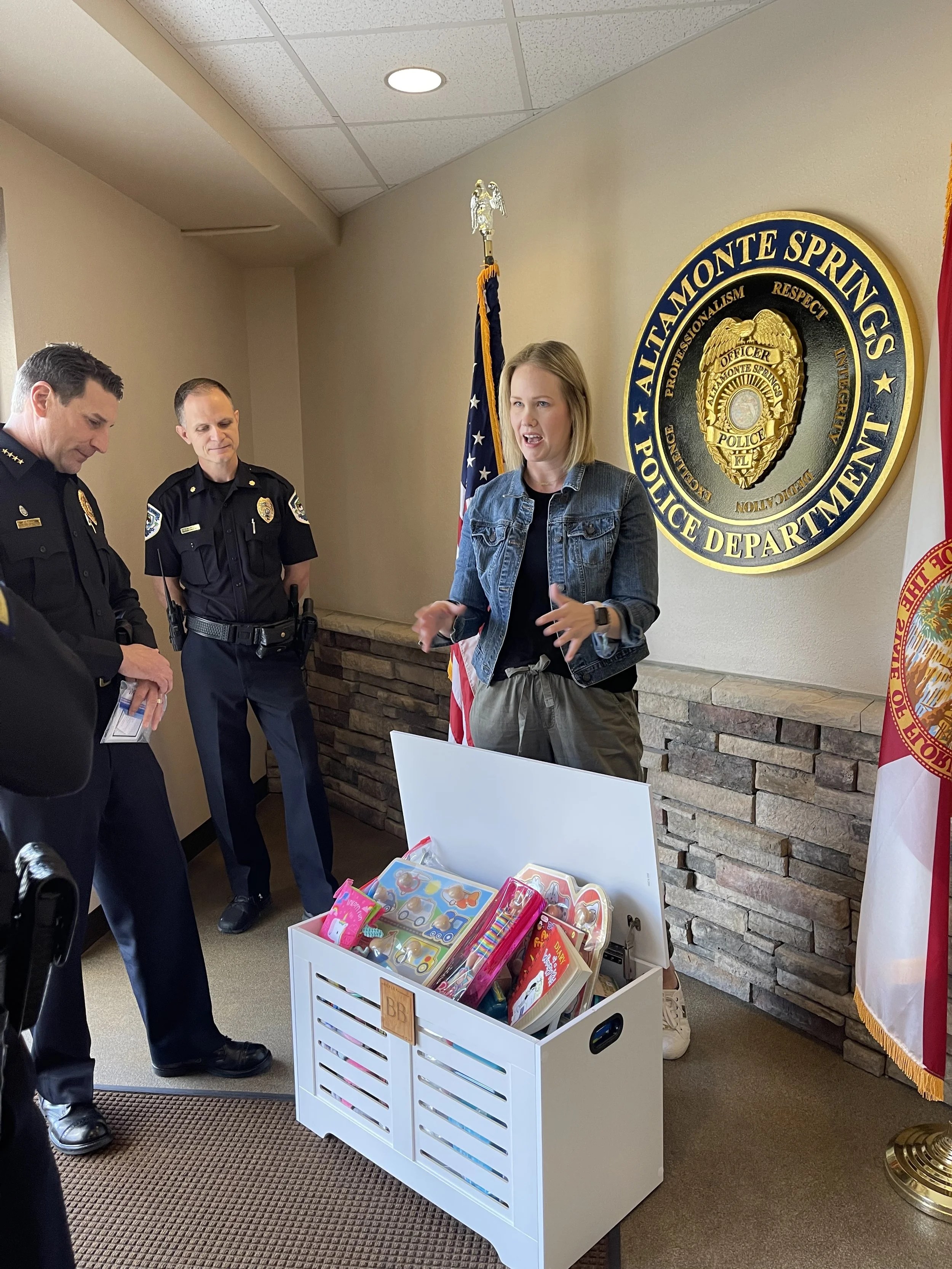 A woman talking to police officers in a room with a sign that reads 'Alamonte Springs Police Department.' She is gesturing with her hands and standing behind a white box filled with toys.