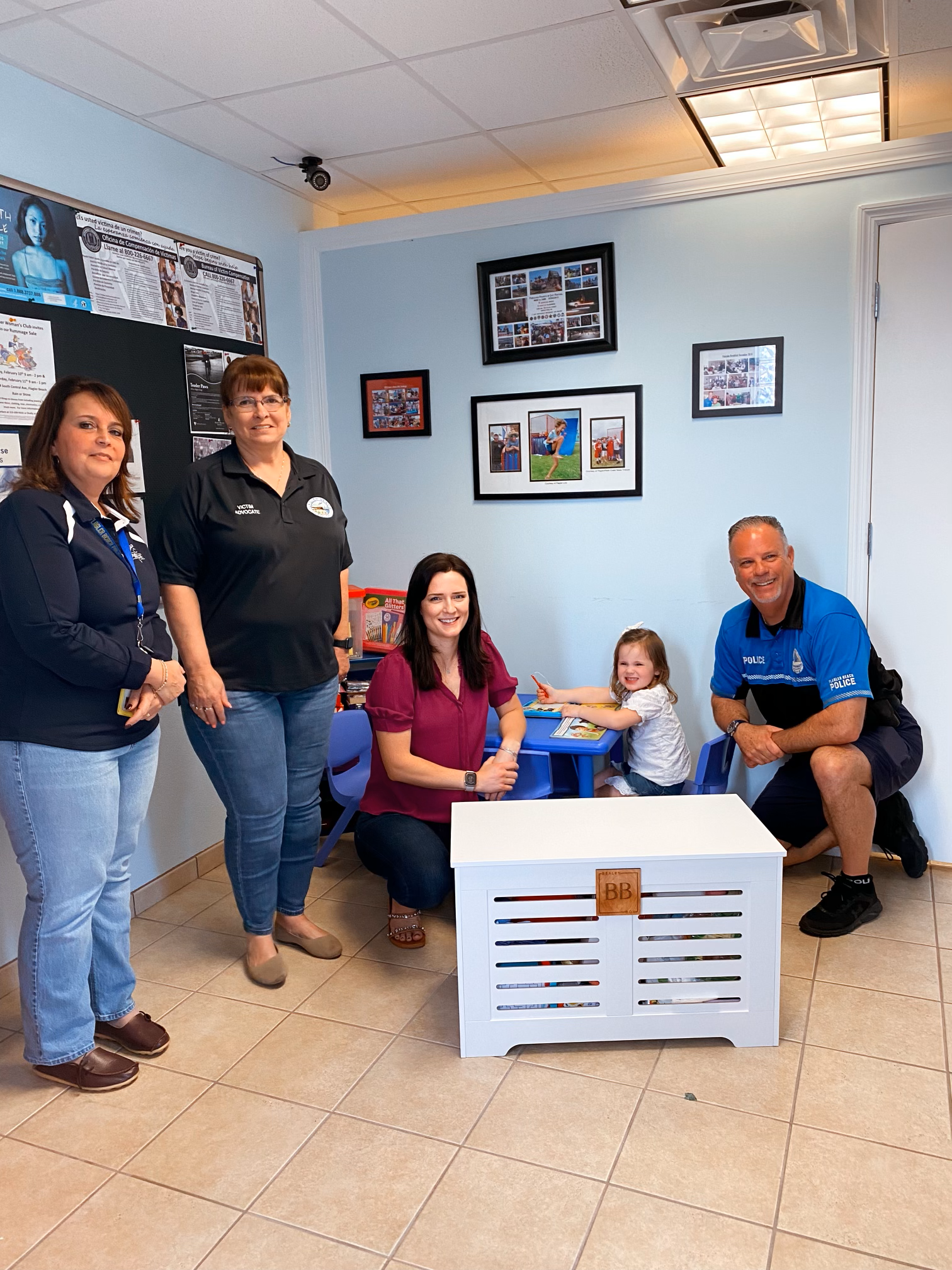 A group of five people, including a young girl, two women, a man in police uniform, and a woman in a black polo shirt, smiling and posing in a small classroom or playroom with blue walls and framed photographs.